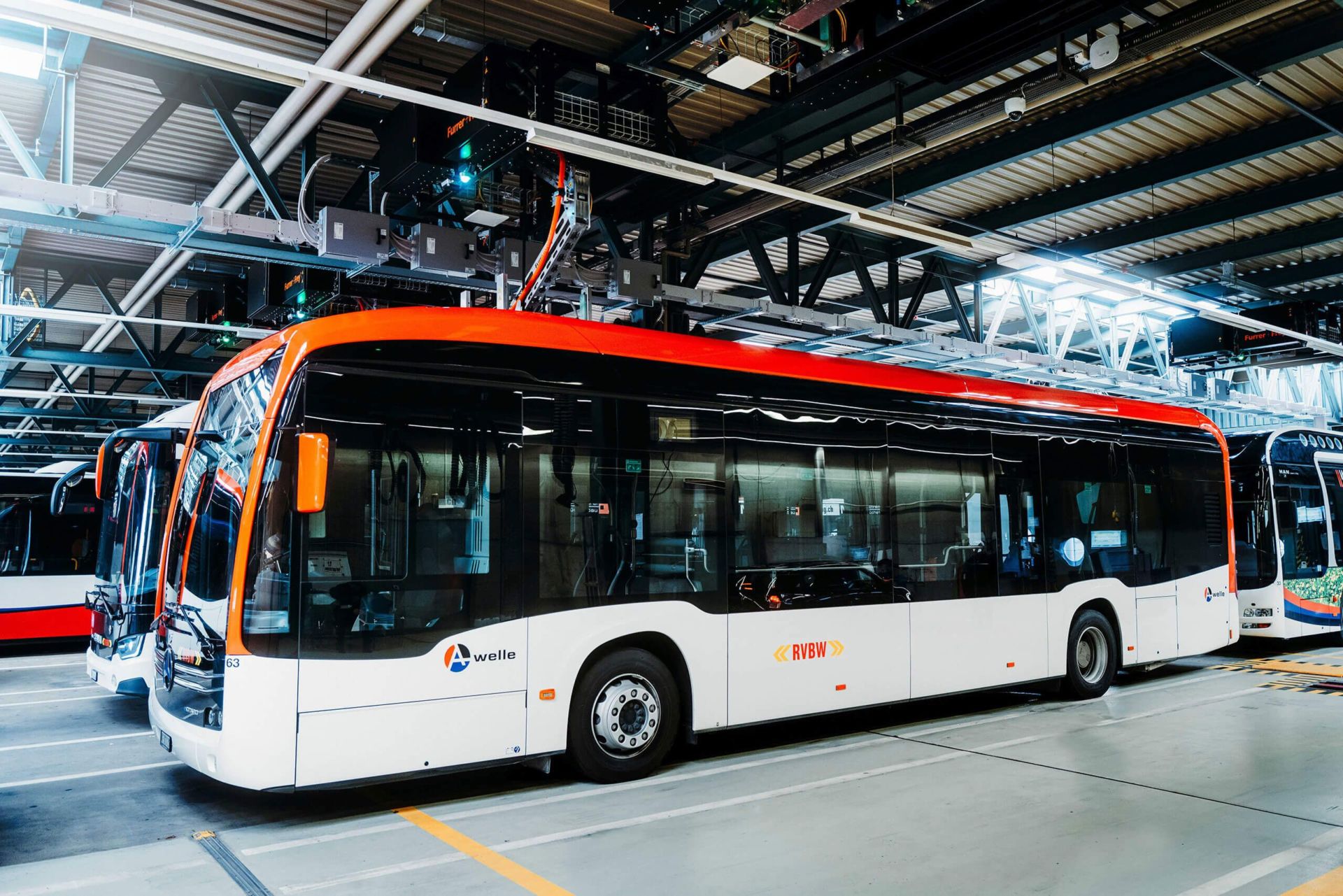 Ein moderner, weißer Elektrobus mit roten und orangen Akzenten steht in einem gut beleuchteten Busdepot. Der Bus mit den Logos „Yvelines“ und „RIVI“ wird von anderen ähnlichen Bussen flankiert. Über ihm ist eine industrielle Anlage mit Geräten und Beleuchtung sichtbar.