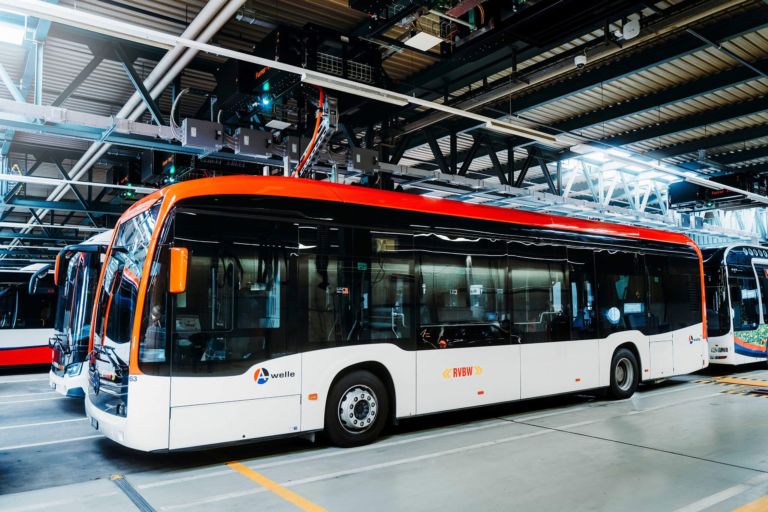 Ein moderner, weißer Elektrobus mit roten und orangen Akzenten steht in einem gut beleuchteten Busdepot. Der Bus mit den Logos „Yvelines“ und „RIVI“ wird von anderen ähnlichen Bussen flankiert. Über ihm ist eine industrielle Anlage mit Geräten und Beleuchtung sichtbar.