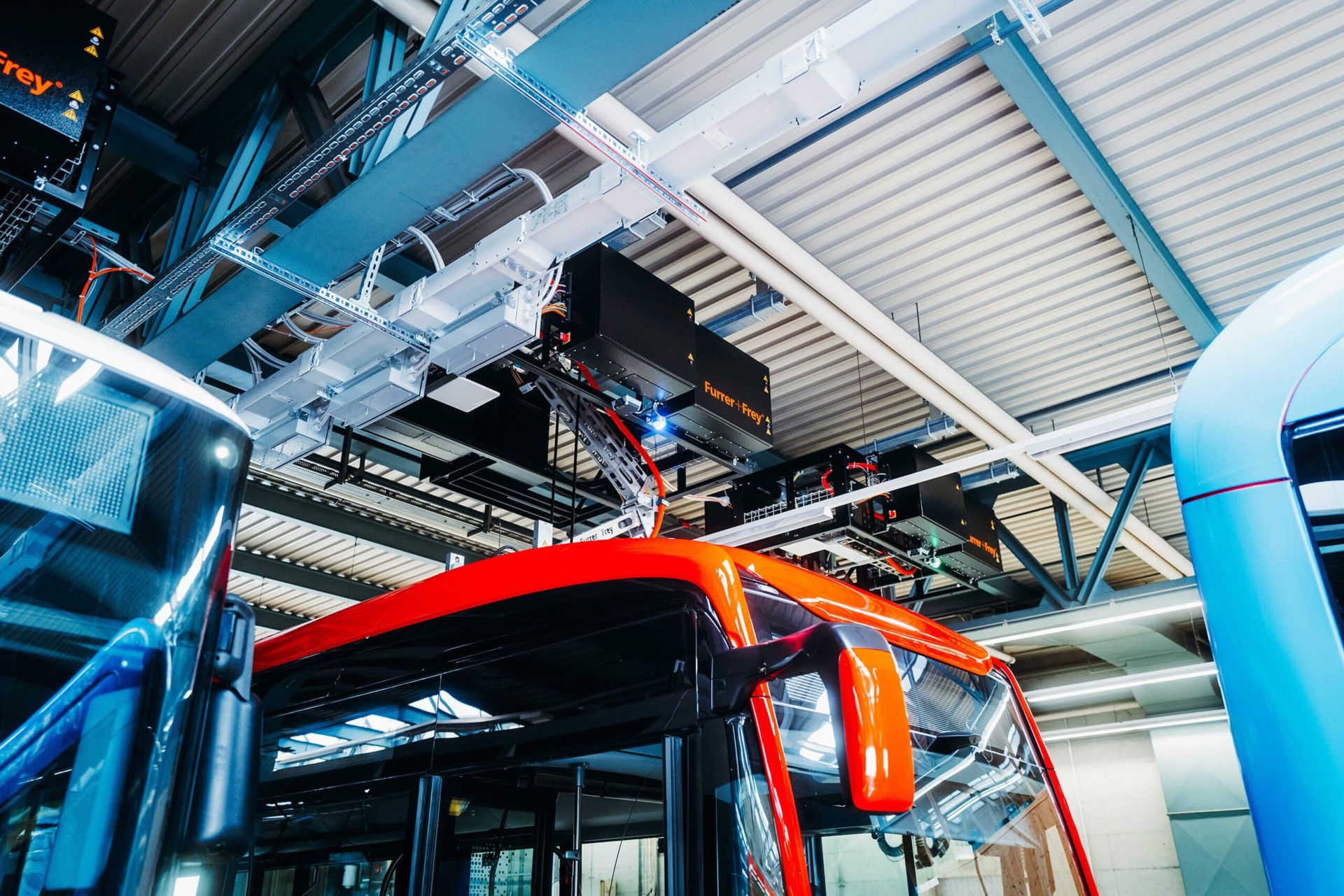 In einer modernen Anlage mit sichtbaren Metallträgern und Industriebeleuchtung wird ein roter Elektrobus über ein Überkopf-Ladesystem aufgeladen. Das Ladegerät ist über dem Bus ausgerichtet und mit einer Struktur an der Decke verbunden. Andere Busse sind teilweise sichtbar.