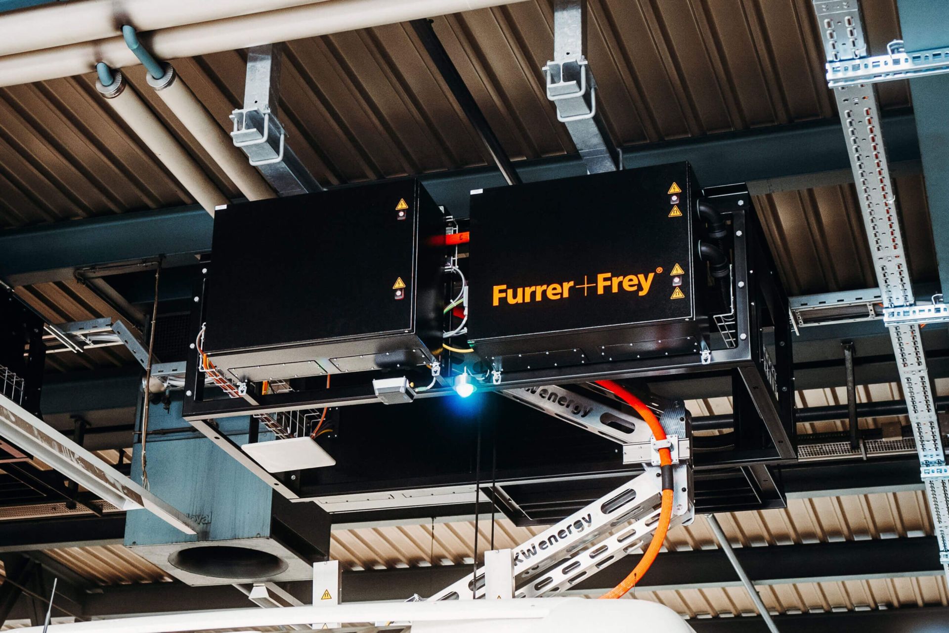 Nahaufnahme eines elektrischen Kopplers von Furrer + Frey, der an der Decke einer Industrieanlage montiert ist. An dem Koppler sind Kabel und verschiedene Warnschilder angebracht. Im Hintergrund sind Metallträger, Rohre und Leitungen zu sehen.