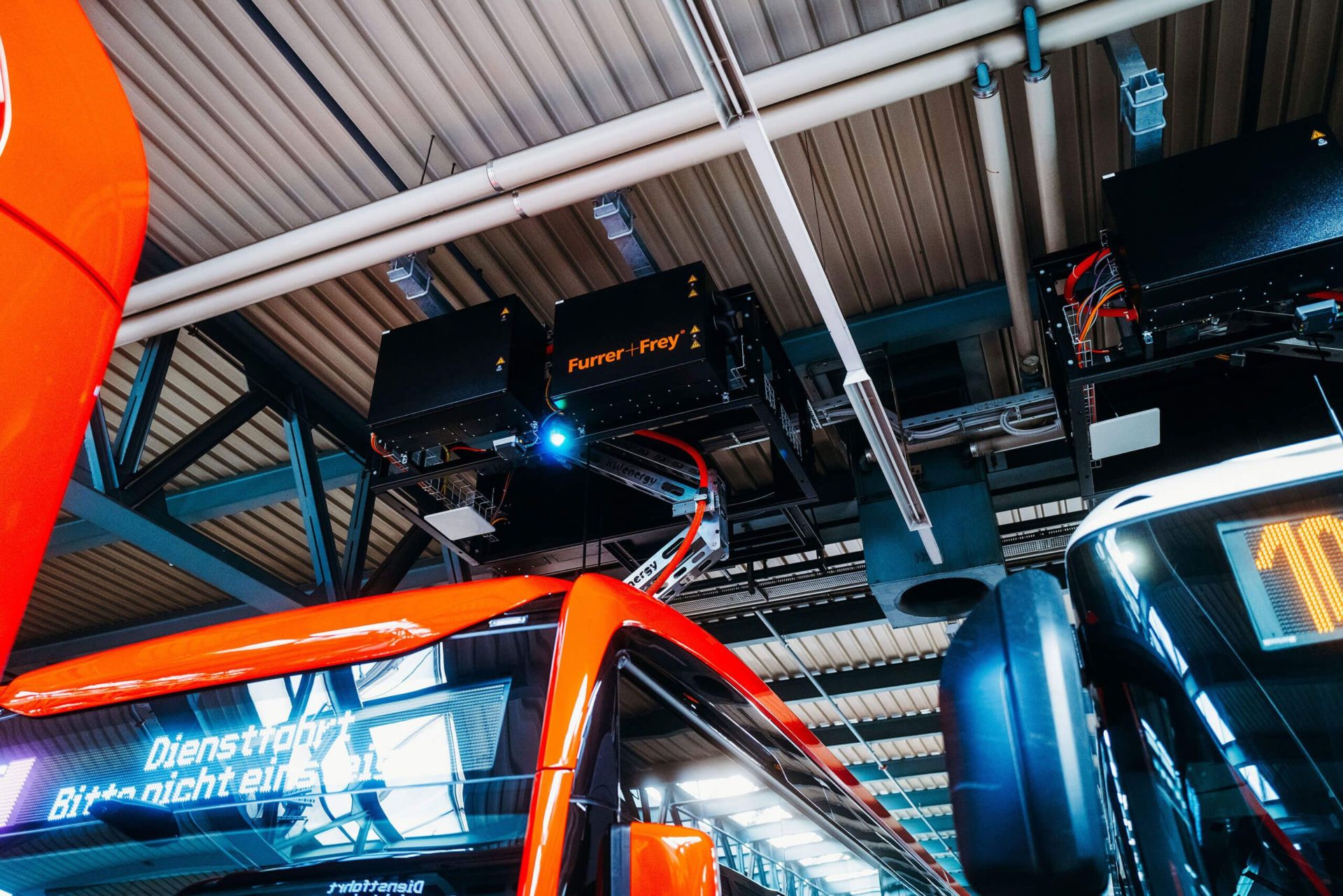 Ein Depot mit Elektrobussen, die über eine Ladeinfrastruktur an der Decke aufgeladen werden. Die Umgebung ist industriell mit Metallträgern und einer Wellblechdecke. Auf der Zielanzeige eines Busses steht „Dienstfahrt“.