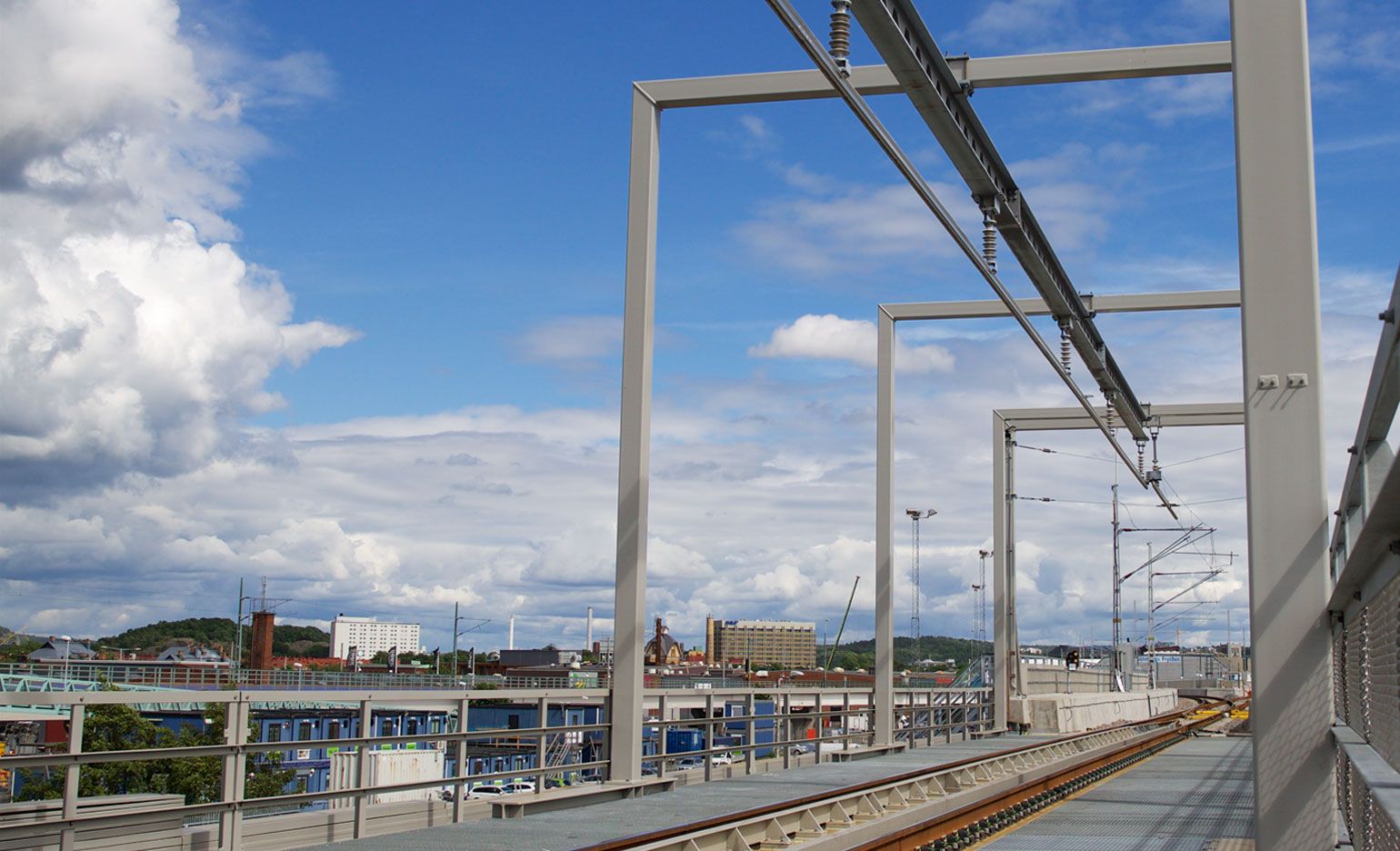 Blick auf eine Hochbahn oder Straßenbahnlinie mit metallischen Überkopfkonstruktionen vor dem Hintergrund einer Stadtlandschaft mit Gebäuden und grünen Hügeln. Der Himmel ist teilweise bewölkt, mit Flecken aus blauen und weißen Wolken. In der Ferne erstrecken sich Schienen und Stromleitungen.