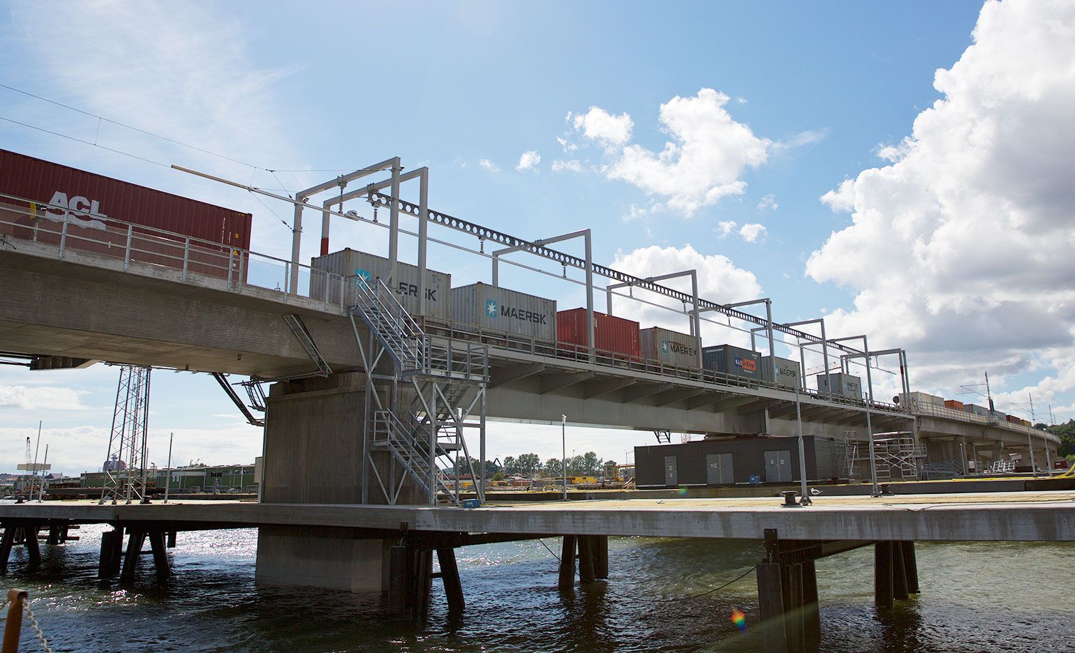 An einem sonnigen Tag befindet sich eine Brücke mit mehreren Frachtcontainern auf Eisenbahnwaggons über einem Gewässer. Die Container sind mit Markennamen beschriftet und die Szene umfasst industrielle Infrastruktur und einen teilweise bewölkten Himmel.