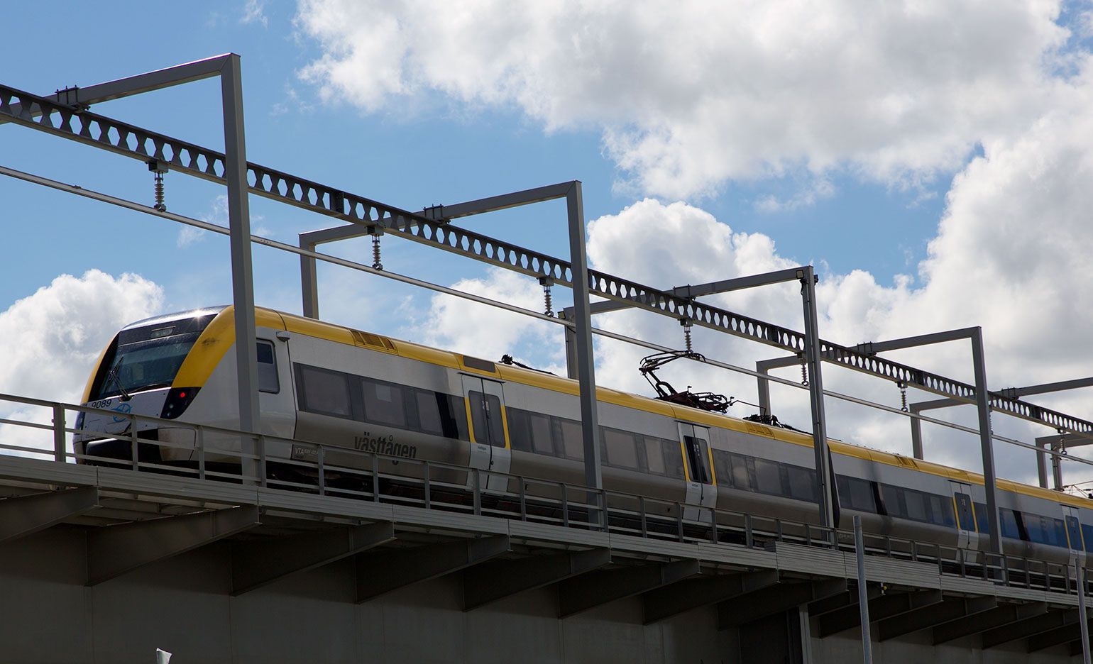 Ein moderner gelb-weißer Zug fährt auf einer Hochbahnstrecke vor einem teilweise bewölkten Himmel. Der Zug wird von Oberleitungen angetrieben, die von Metallportalen getragen werden.