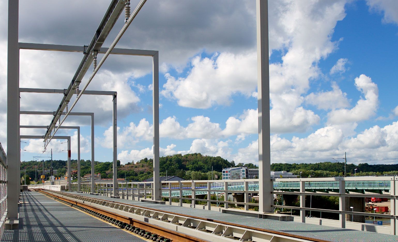 Blick auf eine Bahnstrecke und sich in die Ferne erstreckende Hochstrukturen, mit einem üppig grünen Hügel und verstreuten Gebäuden im Hintergrund unter einem teilweise bewölkten Himmel.