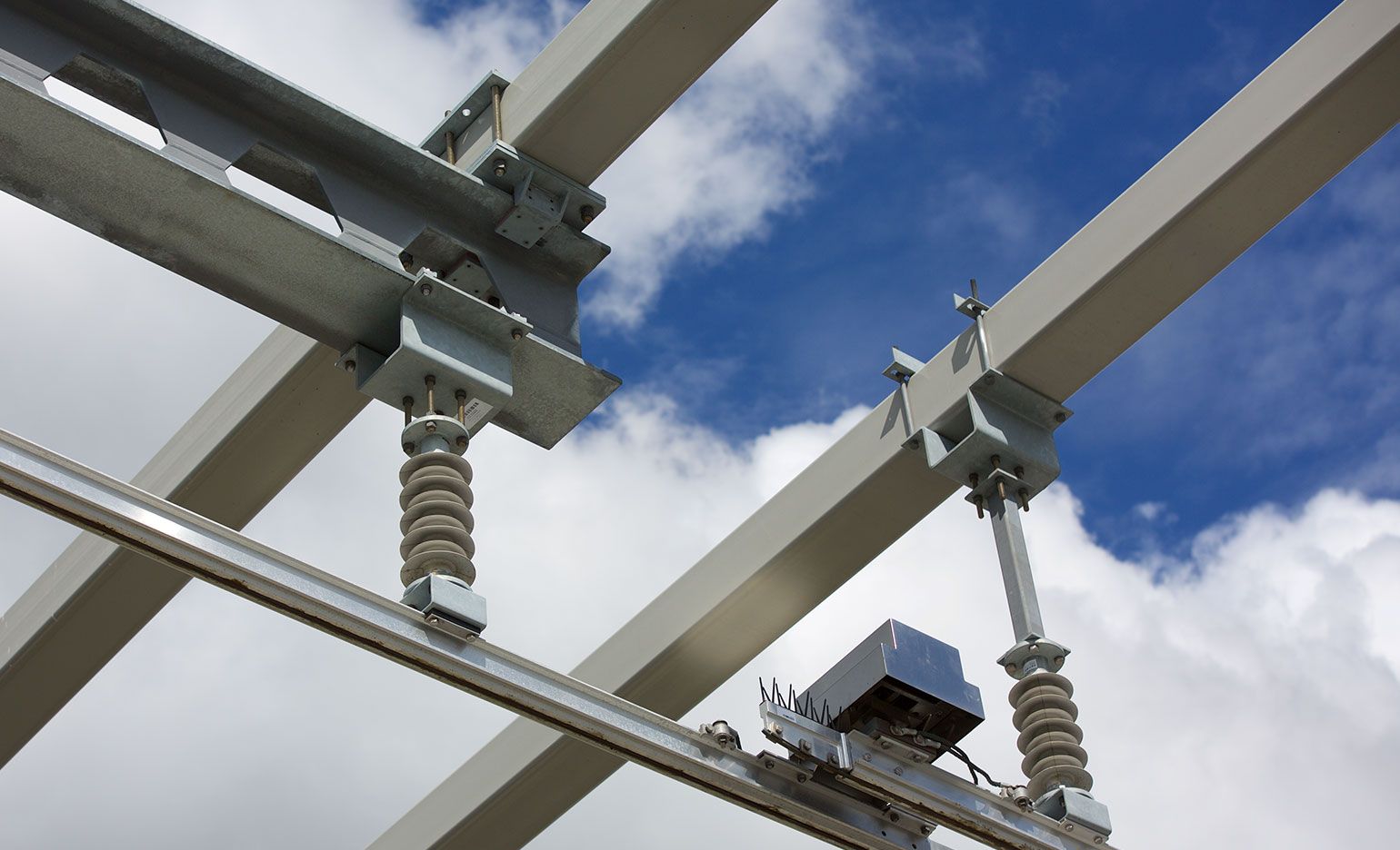 Stahlträger mit Hochleistungsverbindern stehen vor einem strahlend blauen Himmel mit vereinzelten Wolken. Die mit isolierten Stromleitungen und mechanischen Komponenten ausgestatteten Träger lassen auf eine industrielle oder bauliche Umgebung schließen.