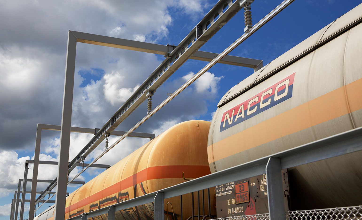 Zwei Kesselwagen, einer orange und einer grau, mit dem NACCO-Logo, stehen auf parallelen Gleisen. Über den Wagen hängen Metallkonstruktionen mit Kabeln und Schläuchen. Die Szene spielt unter einem blauen Himmel mit vereinzelten Wolken.