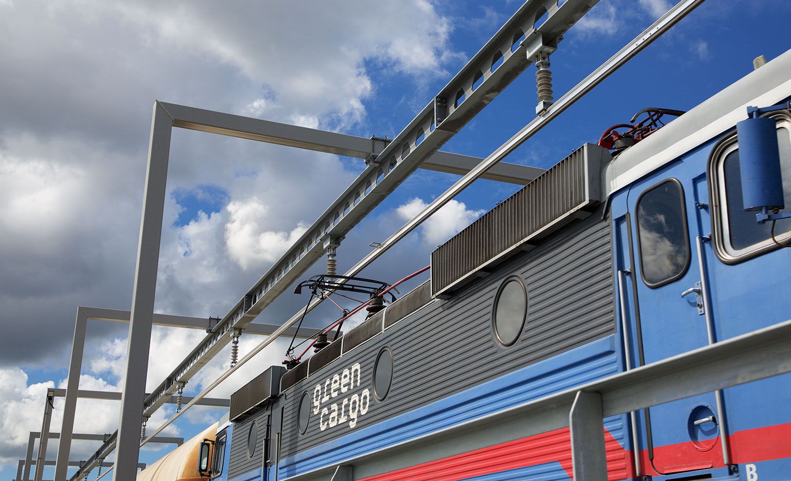 Ein Güterzug mit der Aufschrift „Green Cargo“ auf einem der Waggons fährt auf den Gleisen unter einem teilweise bewölkten Himmel. Der Zug besteht aus verschiedenen Abschnitten in unterschiedlichen Farben, darunter Blau und Rot. Über ihm sind Metallstrukturen zu sehen.
