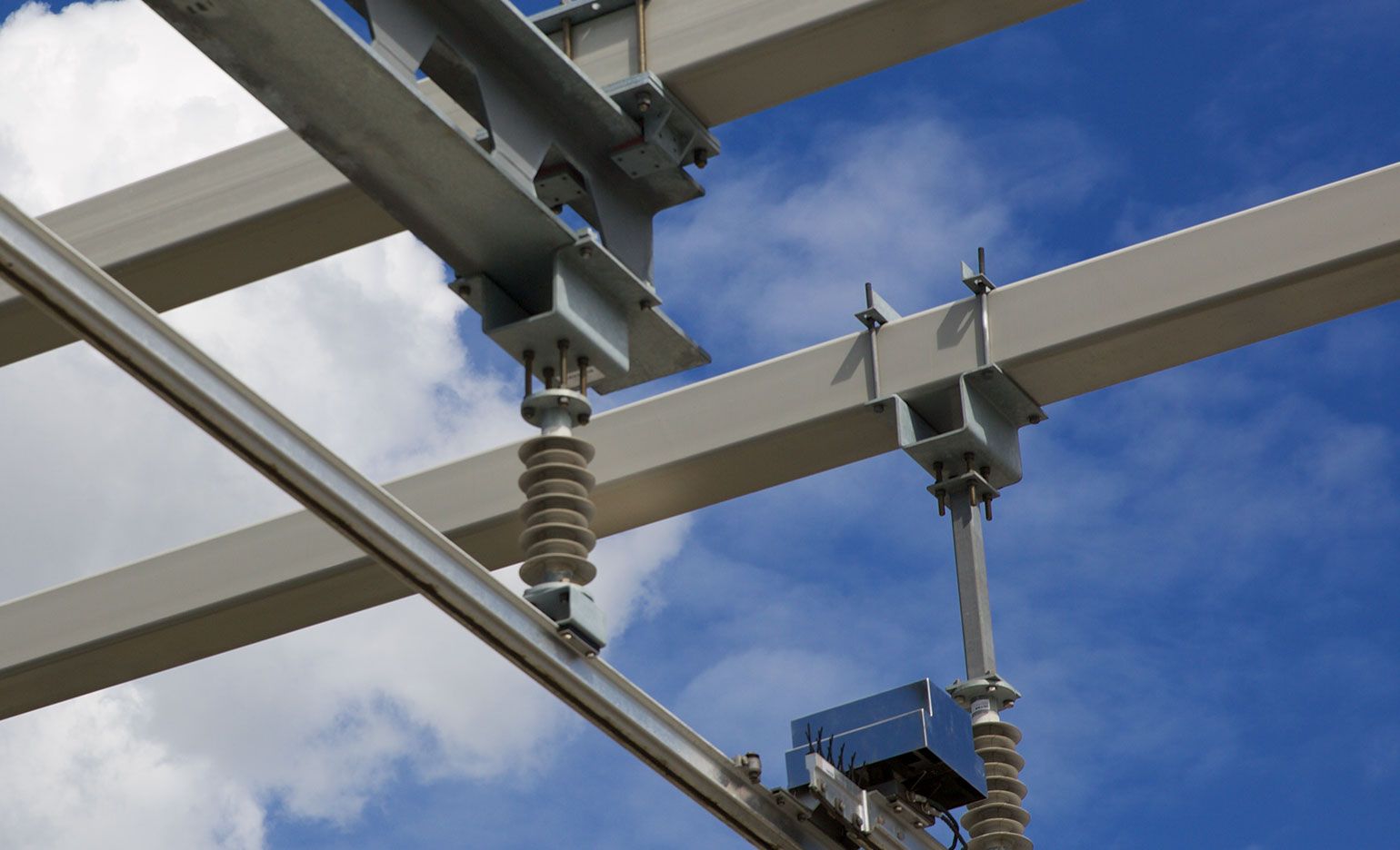 Blick von unten auf die Struktur eines Umspannwerks vor einem strahlend blauen Himmel mit vereinzelten weißen Wolken. Das Bild zeigt Metallträger, Isolatoren und verschiedene Komponenten des Umspannwerksgerüsts.