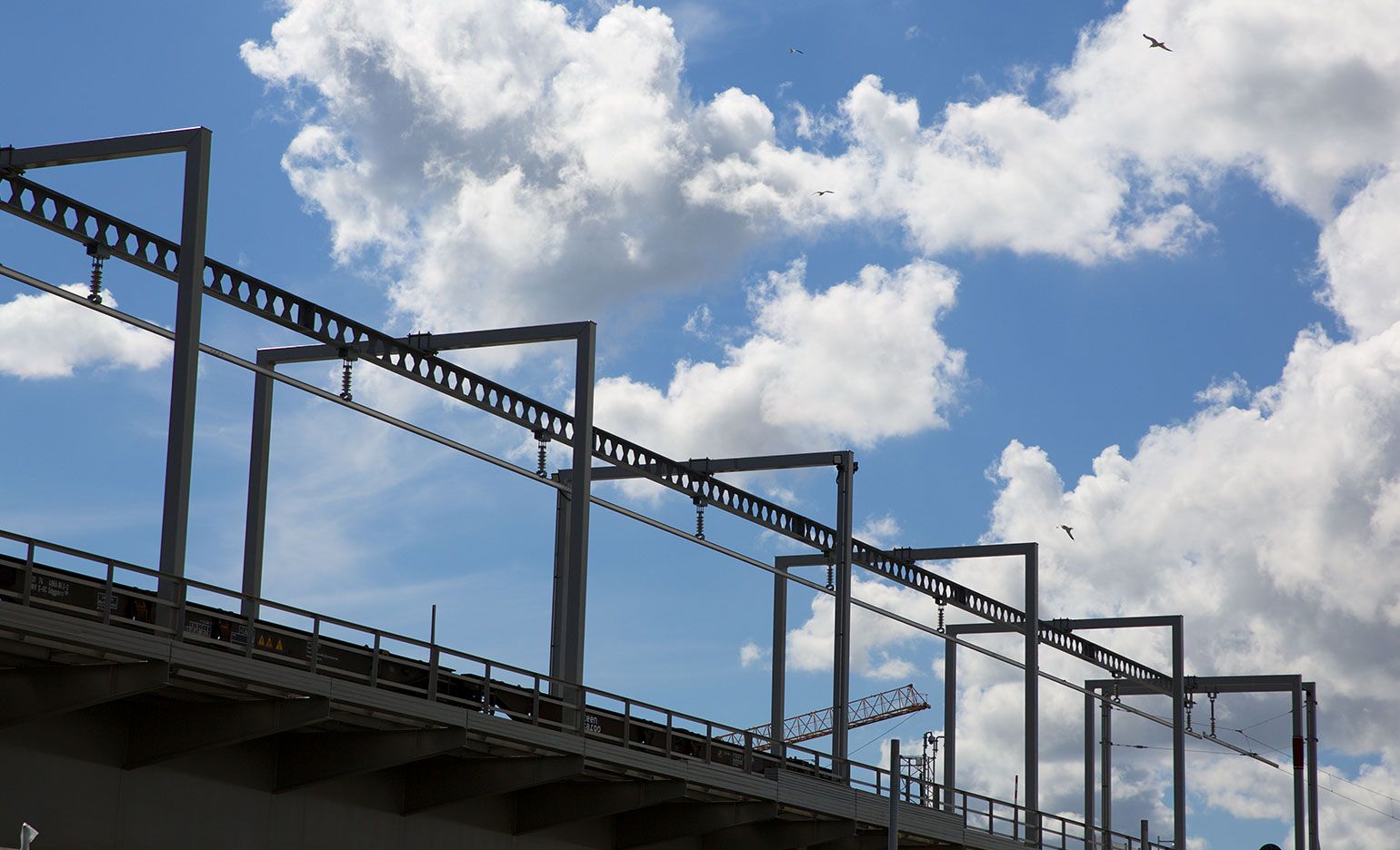 Hinter der Silhouette einer Eisenbahnbrücke mit einer Reihe miteinander verbundener Metallstützstrukturen ist ein blauer Himmel mit vereinzelten Wolken zu sehen. Im Hintergrund fliegen Vögel.
