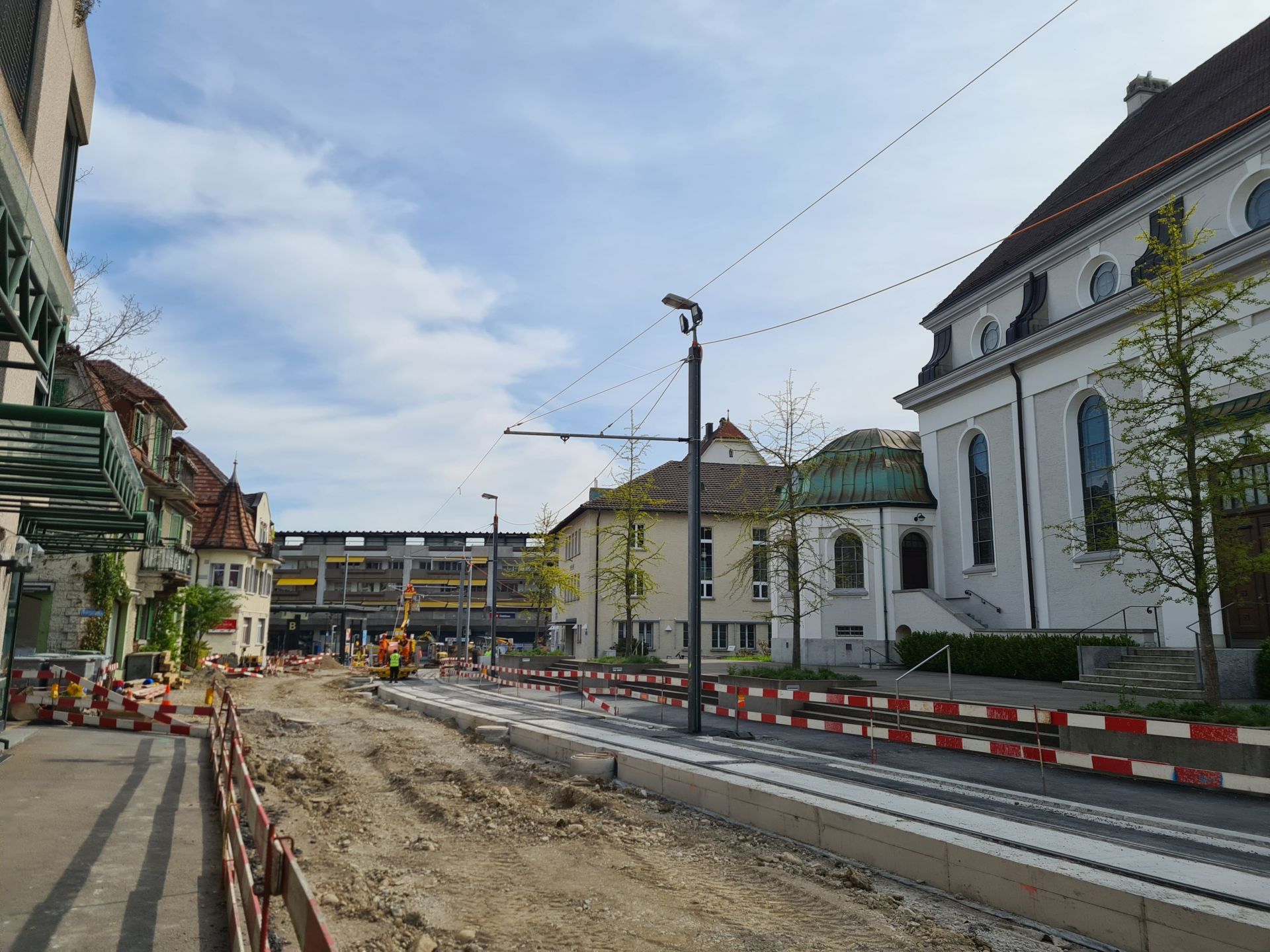Eine Stadtstraße wird gerade gebaut. Der Bereich ist von Absperrungen und Zäunen gesäumt. Am Ende der Straße sind Arbeiter zu sehen. Auf beiden Seiten sind Gebäude mit großen Fenstern und architektonischen Details zu sehen. Oberleitungen und Masten sind vor dem bewölkten Himmel zu sehen.