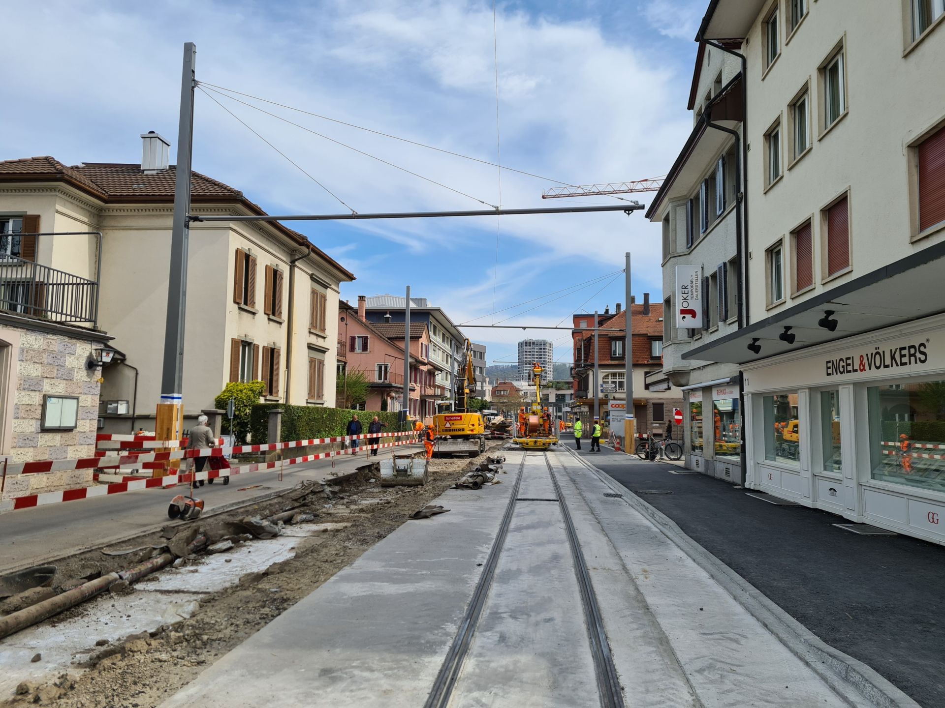 Eine Stadtstraße wird gerade gebaut, in der Mitte werden Gleise verlegt. Auf beiden Seiten der Straße stehen Gebäude, und man sieht Baufahrzeuge und Arbeiter. Ein Fußgängerweg ist abgesperrt, und auf der rechten Seite befindet sich die Ladenfront von Engel & Völkers.
