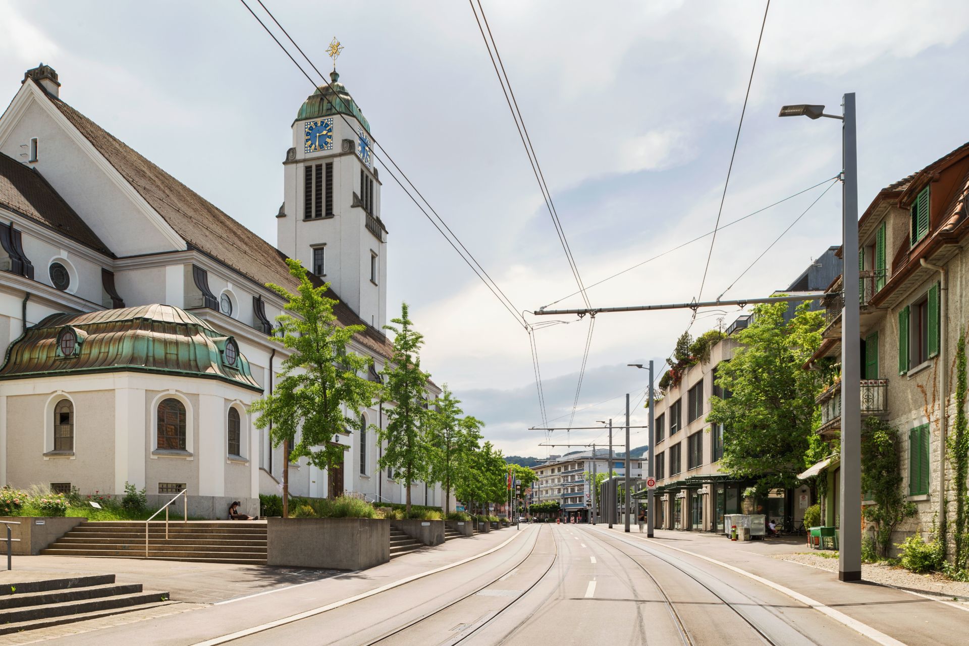 Eine Straßenansicht eines Stadtgebiets mit Straßenbahnschienen, die durch das Zentrum verlaufen. Links ist eine weiße Kirche mit einem Glockenturm und einem grünen Dach. Bäume säumen die Straße, rechts flankieren moderne Gebäude. Der Himmel darüber ist teilweise bewölkt.