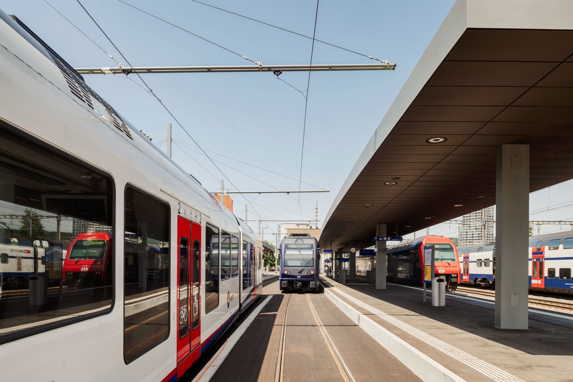 Ein moderner Bahnhof mit mehreren eleganten Zügen in rot-weißen Farbschemata. Der Bahnsteig ist mit einer modernen Dachkonstruktion überdacht und mit elektrischen Oberleitungen ausgestattet, was auf ein elektrifiziertes Schienensystem hindeutet. Der Himmel ist klar und blau.