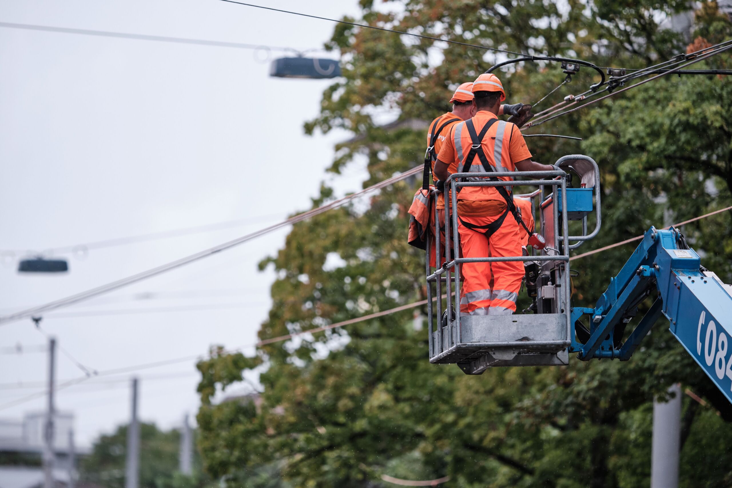 Zwei Arbeiter in orangefarbener Warnkleidung und Helmen reparieren auf einer Hebebühne Oberleitungen. Die Hebebühne ist in der Nähe von Ästen und Stromleitungen angebracht, im Hintergrund ist ein bewölkter Himmel zu sehen.
