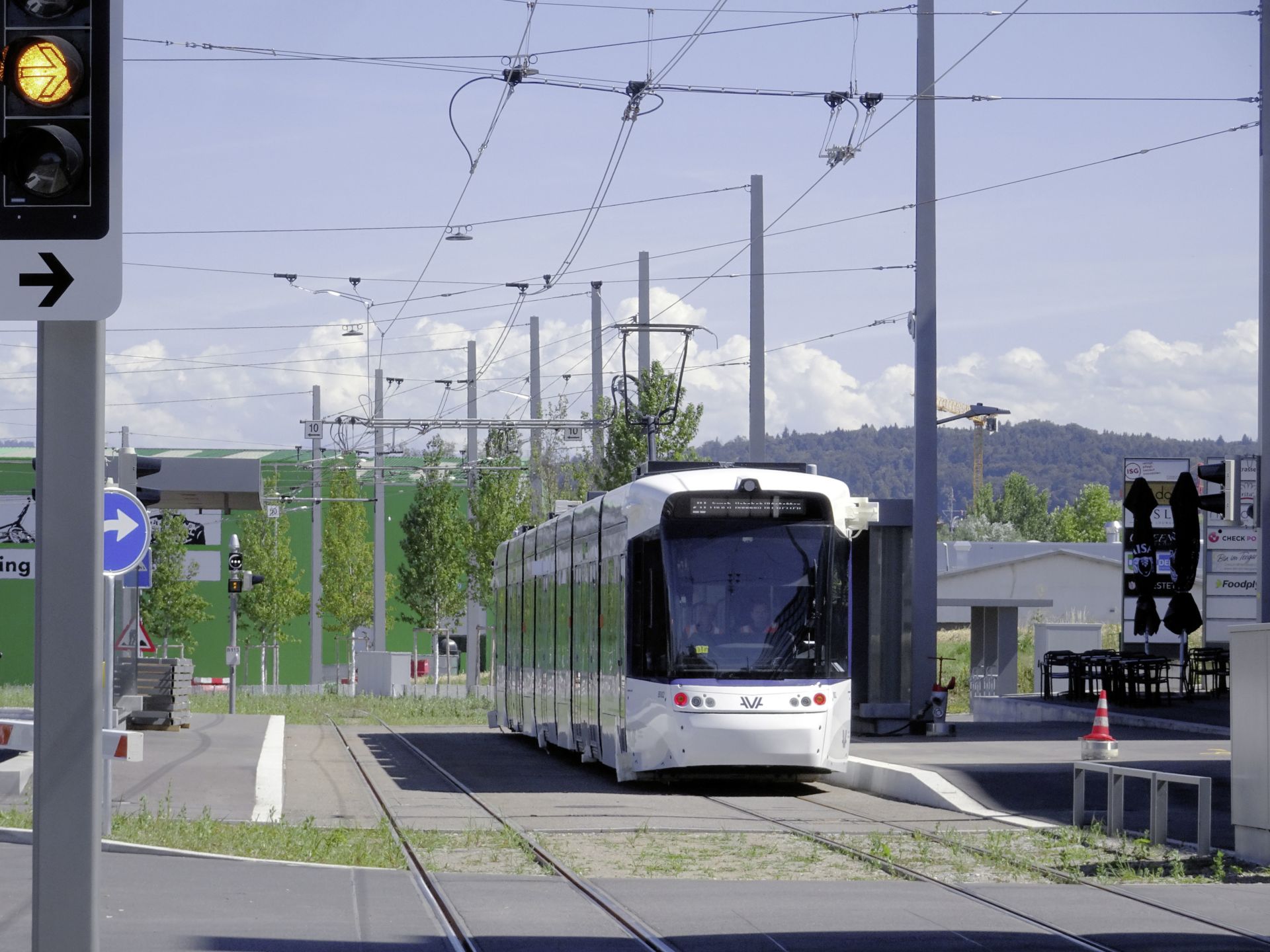 Eine moderne weiße Straßenbahn nähert sich an einem sonnigen Tag einer Haltestelle. Im Hintergrund sind ein grünes Gebäude und bewaldete Hügel zu sehen. Oberleitungen sind sichtbar und auf der linken Seite des Bildes befindet sich eine Ampel. Einige Fahrgäste sitzen in einem Café in der Nähe der Straßenbahnhaltestelle.