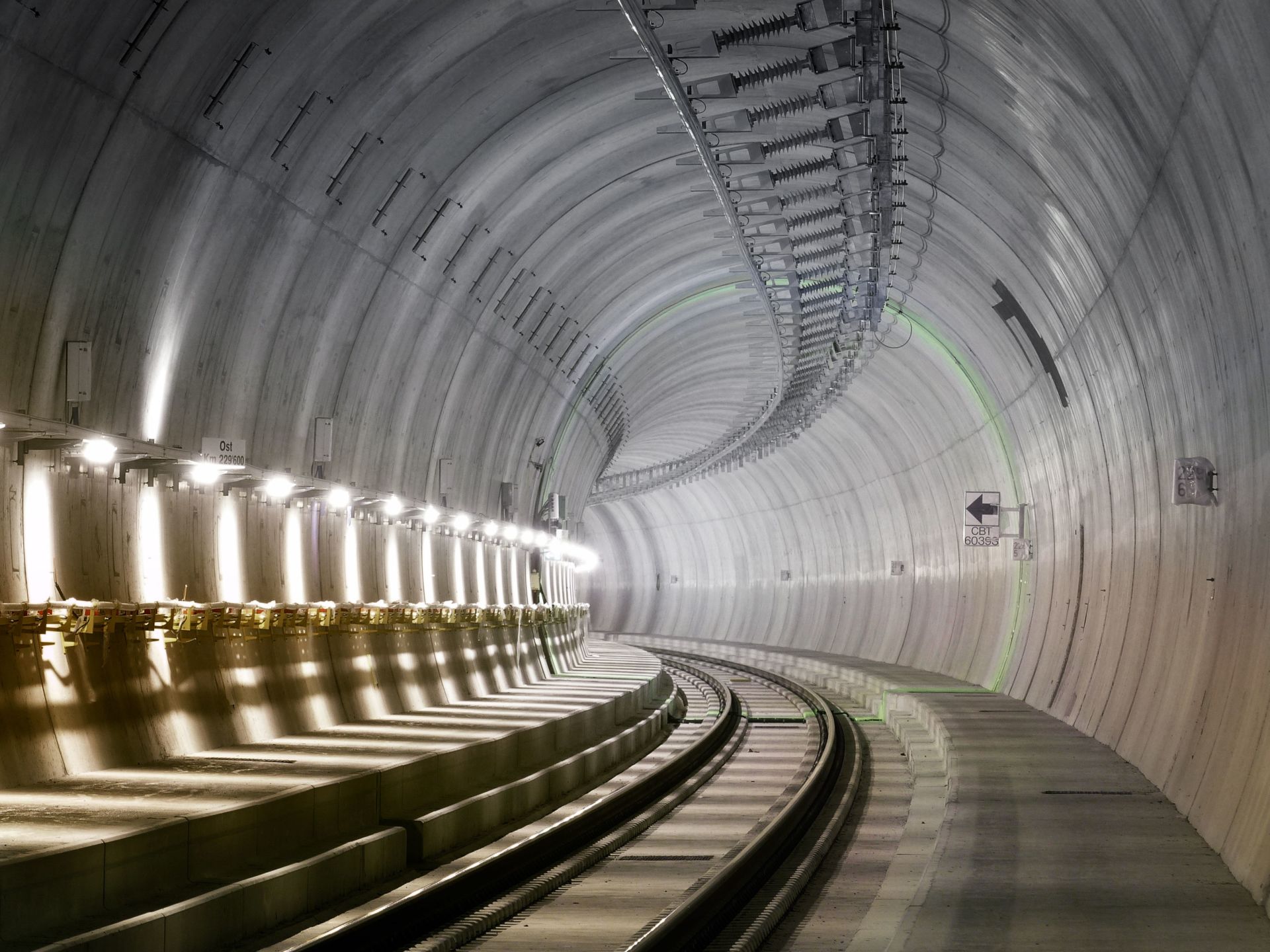 Ein moderner, beleuchteter unterirdischer Tunnel mit geschwungenen Betonwänden. Der Tunnel verfügt über Oberleitungen und zwei nach rechts abknickende Gleise. Symmetrische Leuchten säumen die linke Wand und werfen helles Licht auf die Gleise und den Bahnsteig.