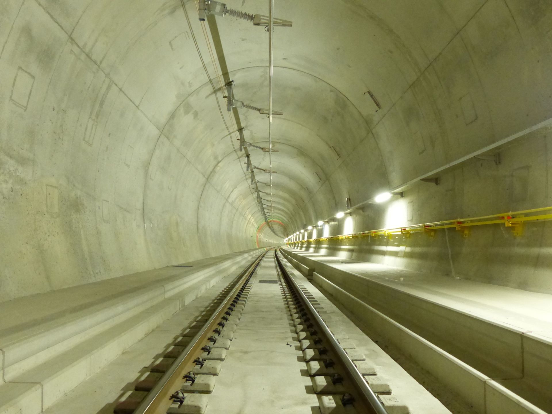 Ein langer Eisenbahntunnel aus Beton mit in die Ferne reichenden Gleisen. Der Tunnel ist durch Deckenleuchten gut beleuchtet und rechts befindet sich ein Fußgängerweg, der mit einem gelben Handlauf gekennzeichnet ist. Die Umgebung wirkt sauber und neu.