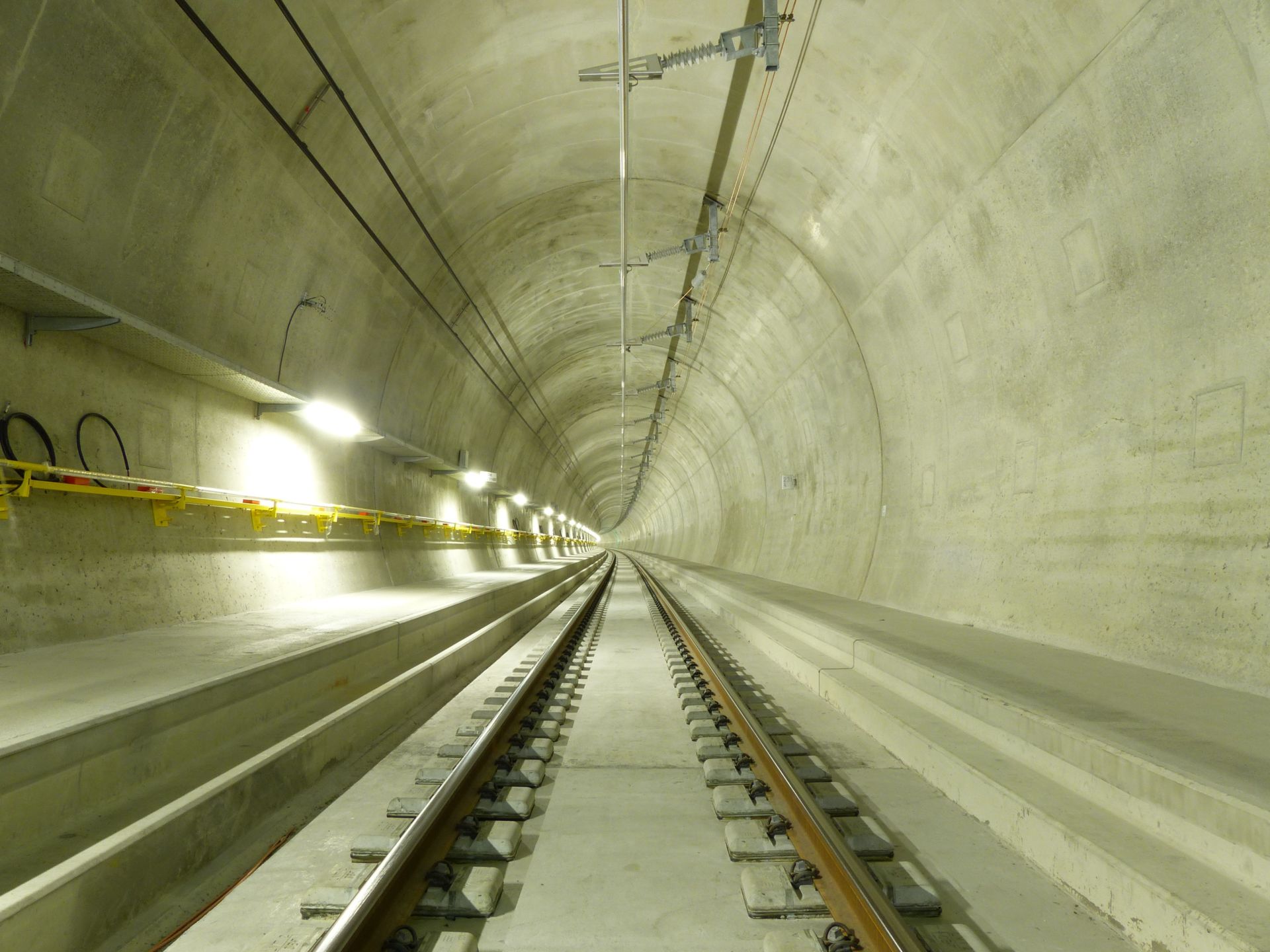 Ein hell erleuchteter Eisenbahntunnel aus Beton erstreckt sich in die Ferne. In der Mitte verlaufen Stahlschienen, die auf beiden Seiten von Betonplatten eingerahmt werden. Stromleitungen und Wartungsgänge sind an den Wänden befestigt und werden von Deckenlampen beleuchtet.