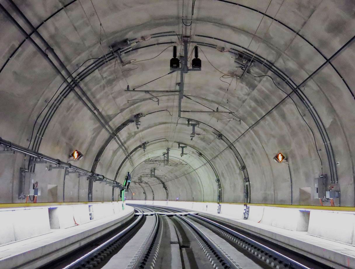 Ein moderner, gut beleuchteter Eisenbahntunnel mit zwei parallelen Gleisen, die leicht nach links abknicken. Der Tunnel hat glatte, gewölbte Betonwände und verschiedene Oberleitungen und Geräte. Die Gleise sind von einer niedrigen Barriere umgeben und an den Wänden sind Sicherheits- oder Notlichter sichtbar.