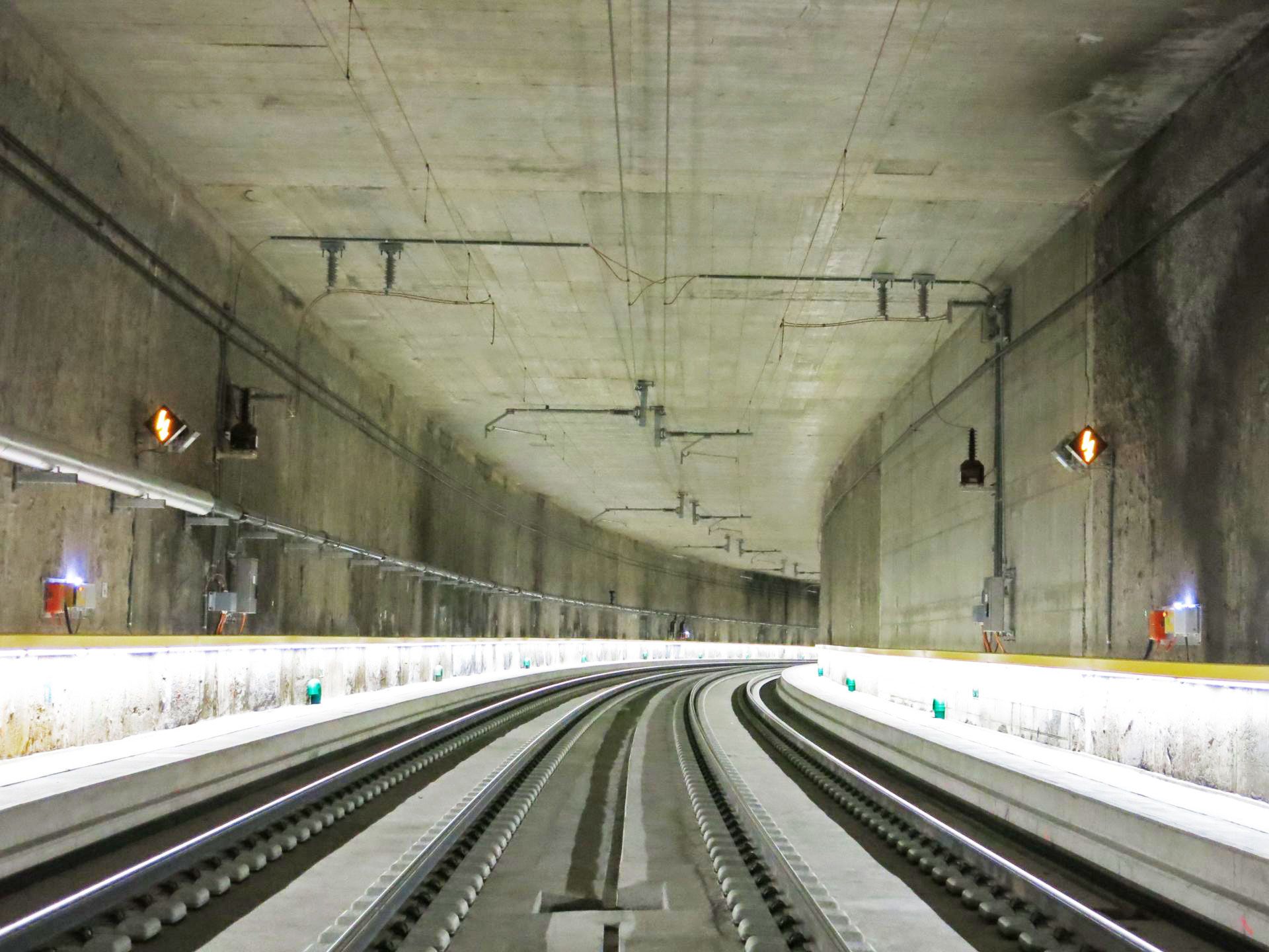 Ein gut beleuchteter, leerer unterirdischer Bahntunnel mit zwei Gleisen erstreckt sich in die Ferne. Die Betonwände und die Decke weisen sichtbare Versorgungsleitungen auf, während sanftes Licht die Gleise und Bahnsteigkanten erhellt.