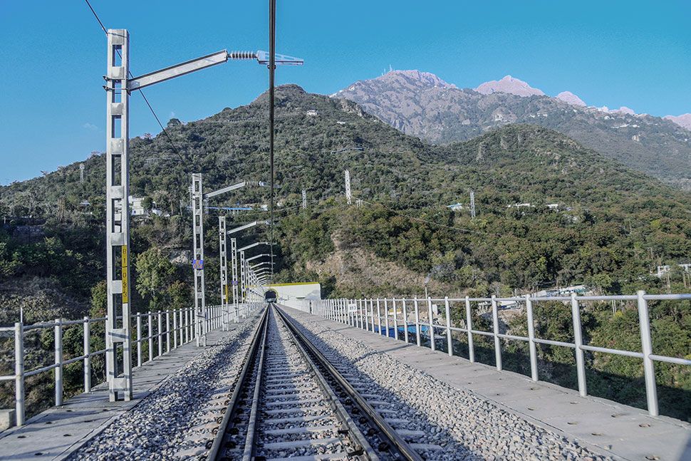 Eine Eisenbahnschienen über eine Brücke, umgeben von grünen Hügeln und Bergen unter einem klaren blauen Himmel. Strommasten säumen die Gleise, und vor uns ist ein Tunnel zu sehen. Es zeigt zudem eine Deckenstromschiene von Furrer+Frey auf offener Strecke auf einer Brücke vor einem Tunnel