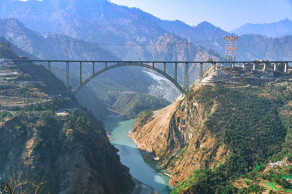 Eine grosse Brücke über einem tiefen Tal mit einem darunter liegenden Fluss, umgeben von schroffen Bergen und grünen Hügeln unter einem klaren Himmel. An einem Ende der Brücke sind Baukräne zu sehen. Die Fahrleitungen über der Brücke sind neu und von Furrer+Frey