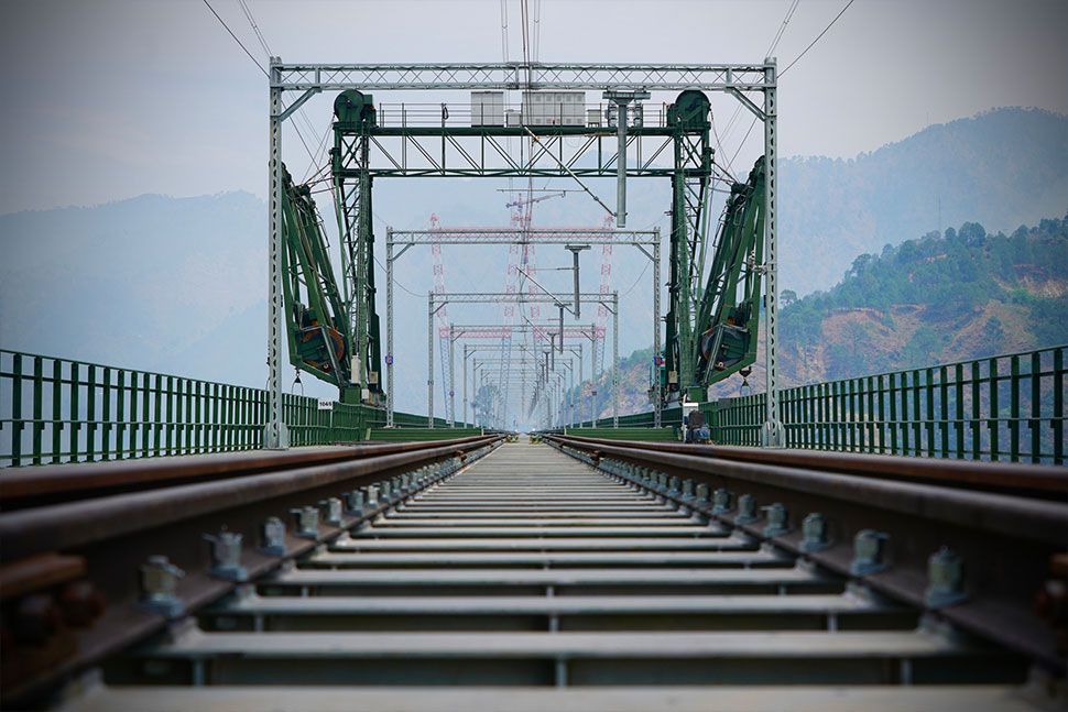 Blick entlang einer Eisenbahnstrecke auf eine grüne Stahlbrücke mit Bergen und Dunst im Hintergrund; die Perspektive betont Symmetrie und Tiefe. Es ist ein Fahrleitungssystem von Furrer+Frey auf der Brücke zu sehen