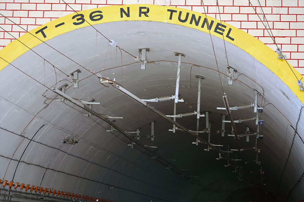 Eingang eines Tunnels mit der Aufschrift "T-36 NR TUNNEL" in schwarzer Schrift auf gelbem Grund. Im Inneren des Tunnels sind elektrische Fahrleitungen und Isolatoren zu sehen. Es handelt sich hierbei um die Deckenstromschiene von Furrer+Frey die an der Tunneldecke montiert sind.
