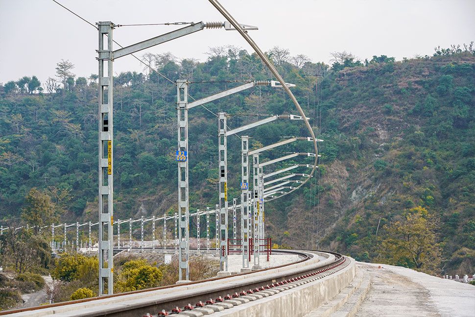 Eine Kurve einer Eisenbahnstrecke mit Fahrleitungen verläuft entlang grüner, baumbewachsener Hügel unter einem bewölkten Himmel. Es zeigt eine Deckenstromschiene von Furrer+Frey auf offener Strecke