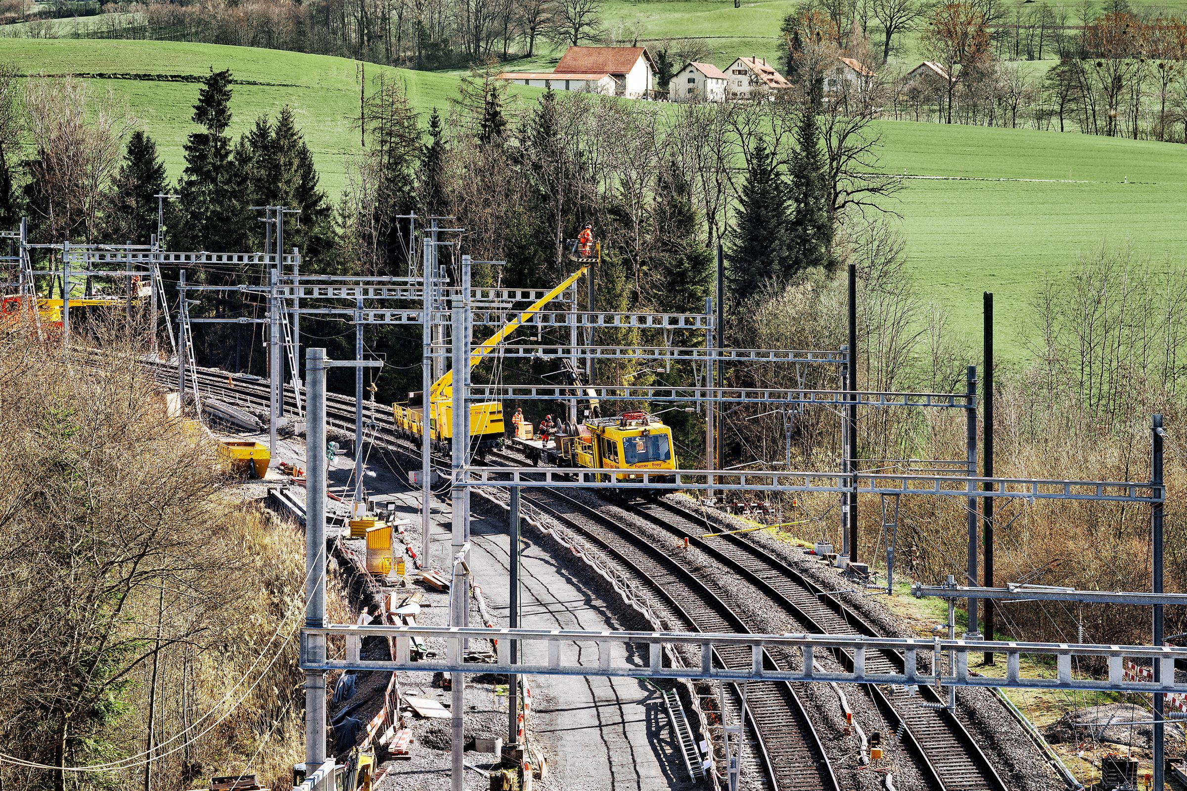 In einer ländlichen Gegend mit grünen Feldern, Bäumen und entfernten Häusern sind Monteure an Fahrleitungsarbeiten der Bahn beschäftigt. Entlang der Gleise verlaufen Strommasten und Leitungen.