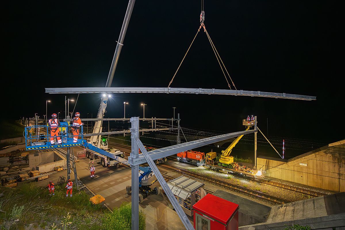 Monteure in orangefarbener Schutzkleidung heben und montieren nachts mit einem Kran eine grosses Joch über einem Bahngleis, wobei Baugeräte und Scheinwerfer die Szene am Tunnelportal beleuchten.