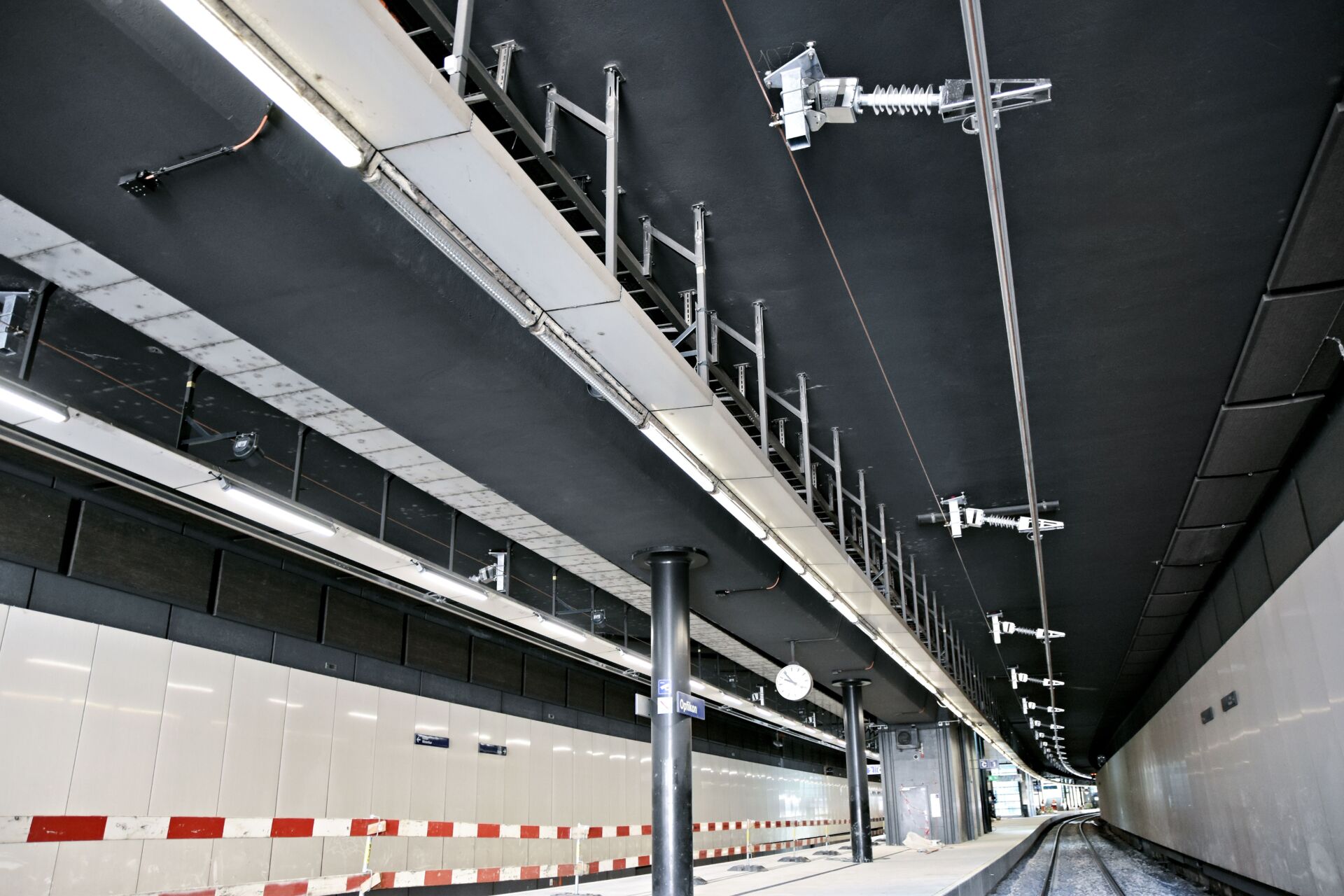Ein moderner Tunnel mit Gleisen, einer weiss gefliesten Wand, Überwachungskameras, Uhren und Deckenstromschiene an der Decke. Der Tunnel ist leer und gut beleuchtet.