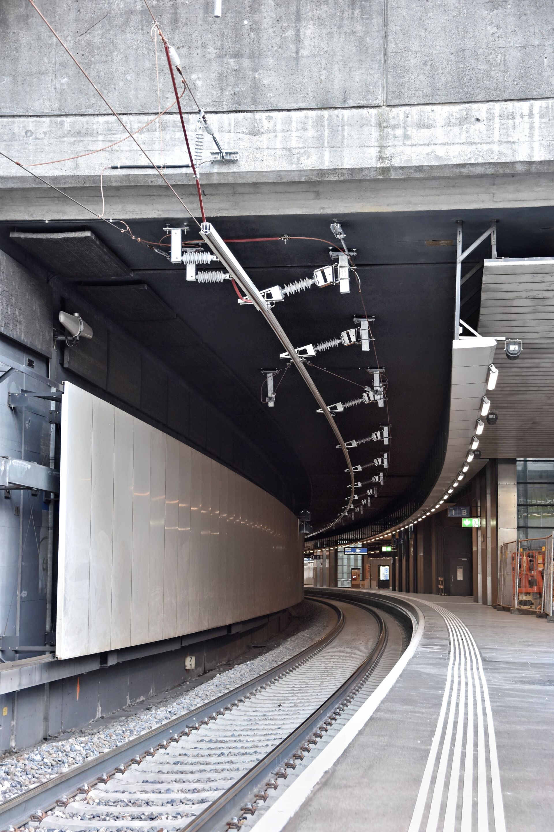 Eine geschwungene Eisenbahnstrecke, Systemübergang Gleis 1, die unter einer modernen städtischen Betonbrücke mit Deckenstromschiene und taktilen Bodenmarkierungen für Barrierefreiheit hindurchführt; an der Decke reihen sich Leuchten aneinander.