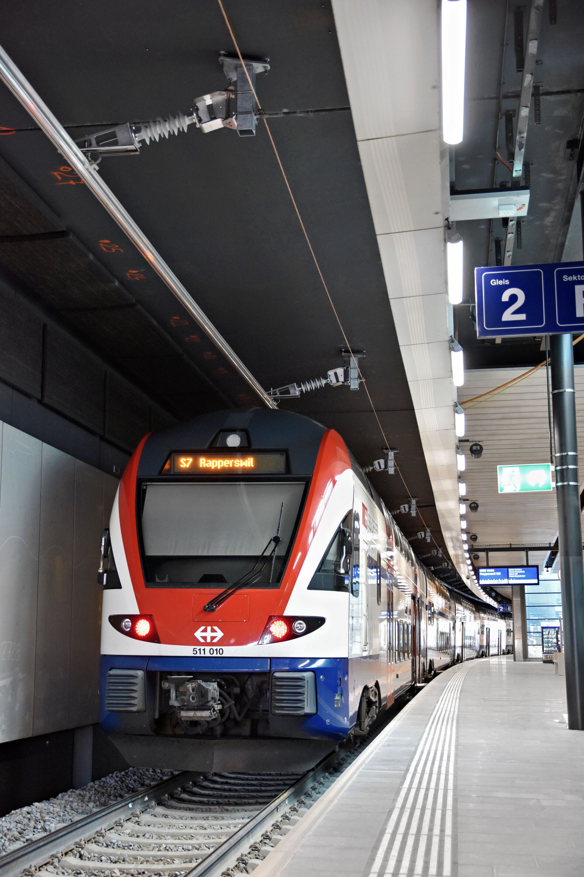 Ein moderner rot-weiß-blauer Zug der SBB mit der Aufschrift „S7 Rapperswil“ steht am Bahnsteig Gleis 2 , wobei die Deckenbeleuchtung und der taktile Bodenbelag zu sehen sind.