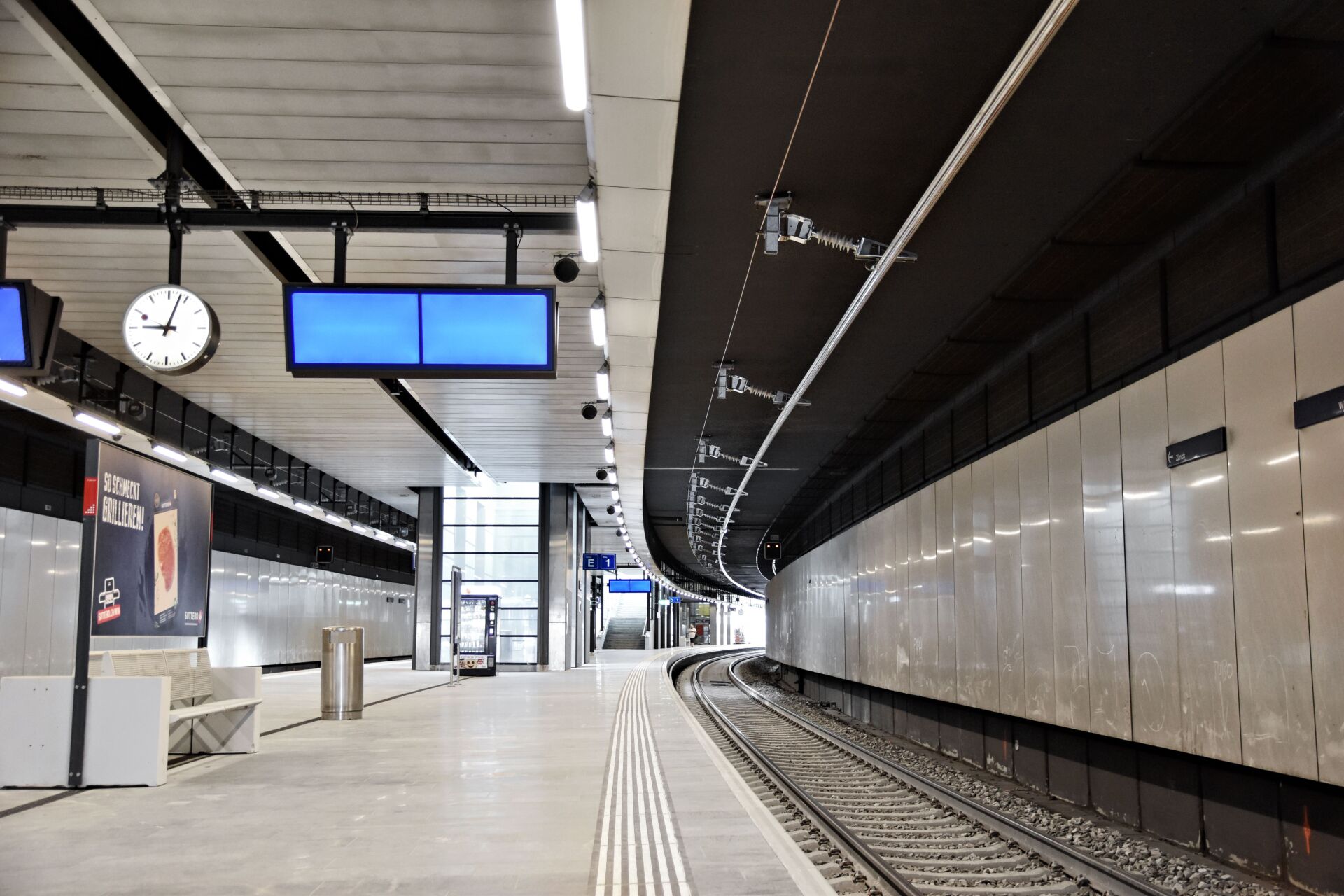Leere Gleise im Bahnhof Opfikon mit Bahnsteig, mit der neuen Deckenstromschiene CR4 und einer Anzeige mit den Bahnsteignummern. Der Bereich ist hell beleuchtet, und es sind keine Menschen zu sehen.