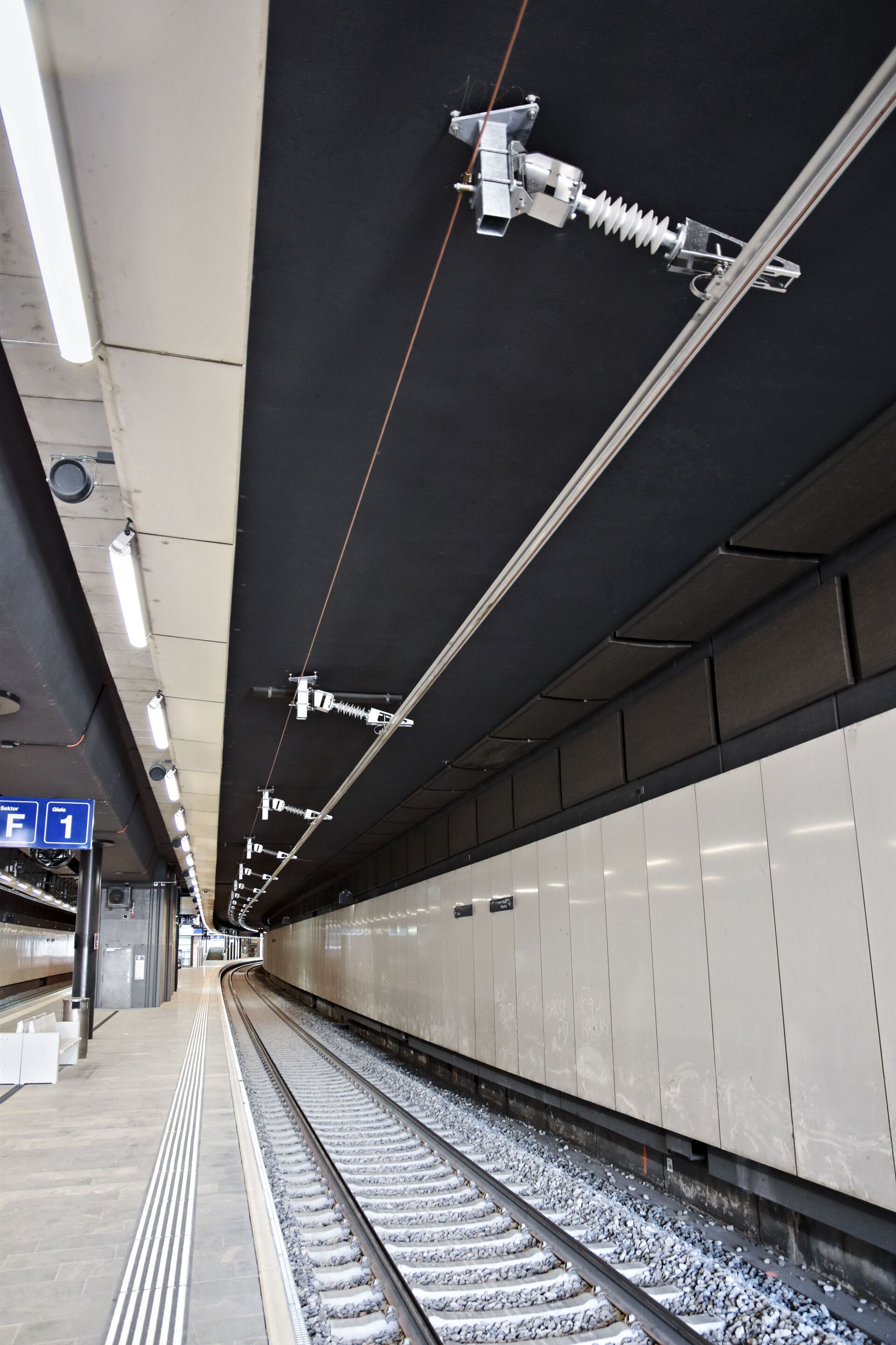 Leere Gleise im Bahnhof Opfikon mit Bahnsteig, mit der neuen Deckenstromschiene CR4 und einer Anzeige mit den Bahnsteignummern. Der Bereich ist hell beleuchtet, und es sind keine Menschen zu sehen.