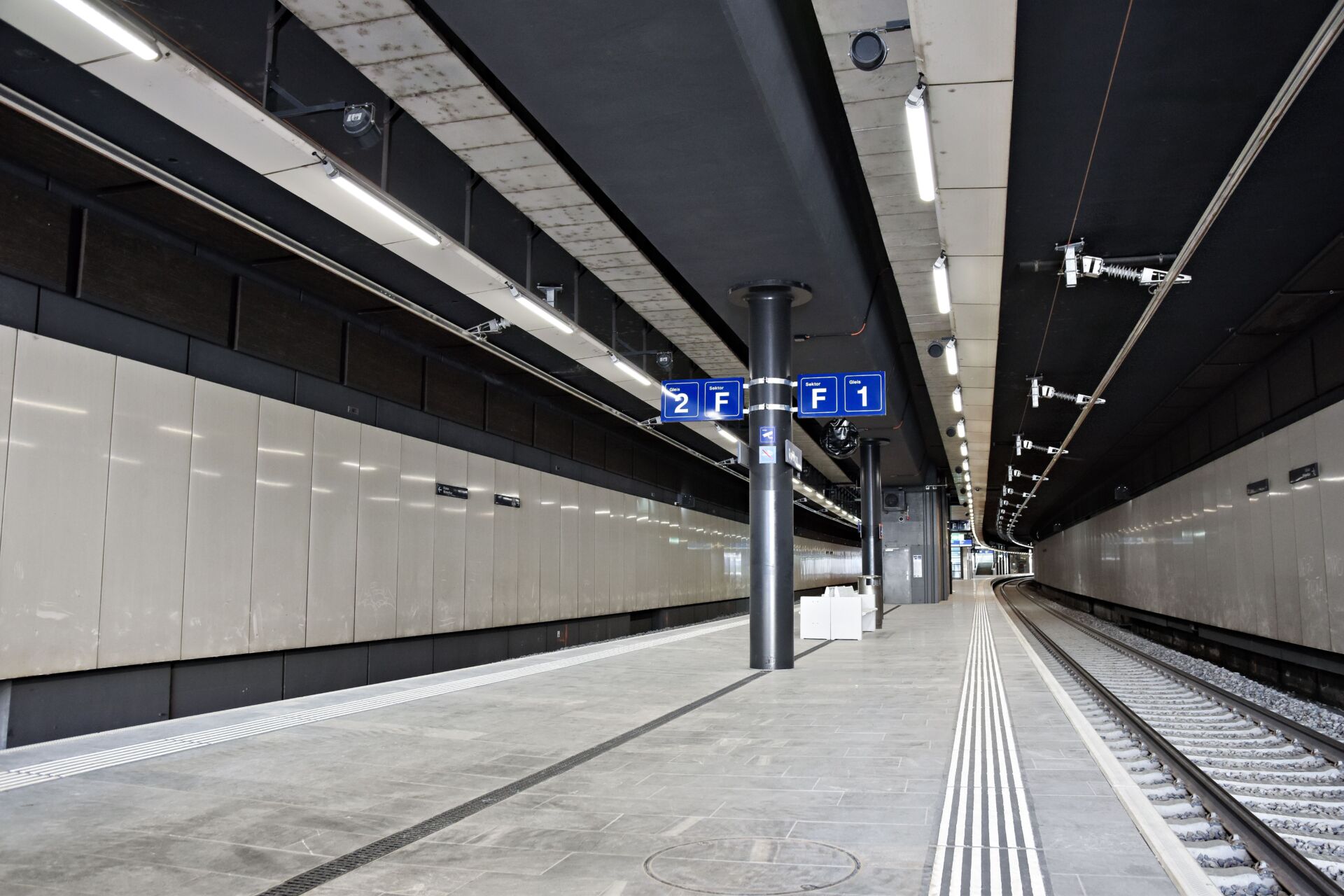 Ein moderner, leerer Bahnsteig am Bahnhof Opfikon mit zwei Gleisen, Überkopfschildern mit den Aufschriften „2 F“ und „1 F“, einer neuen Deckenstromschiene CR4 sowie einem taktilen Bodenbelag am Rand. Der Bahnhof ist sauber, mit beigen Wänden und einer dunklen Decke.