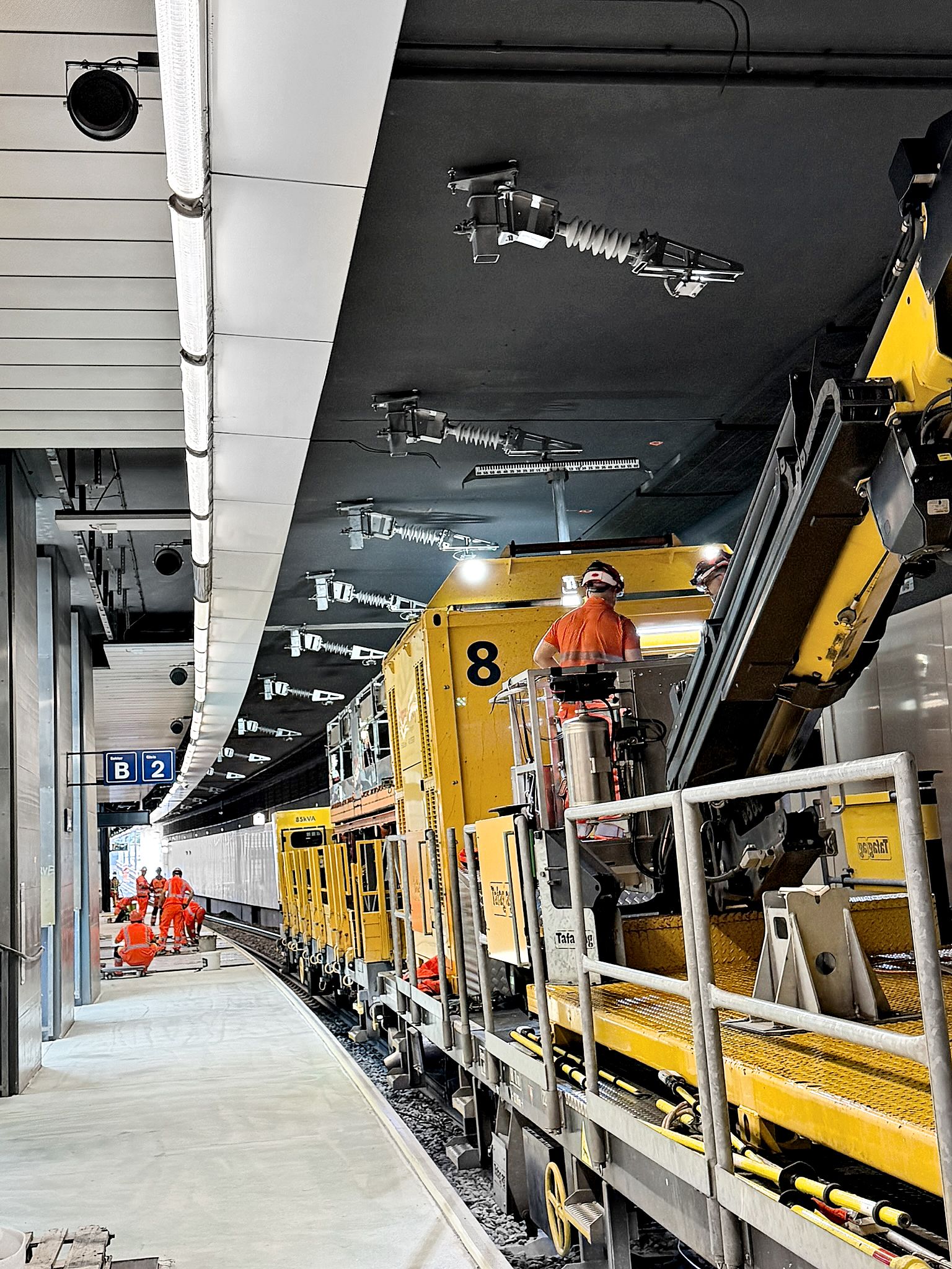 Arbeiter in orangefarbener Sicherheitskleidung bedienen den gleben Montagezug auf den Gleisen im modernen, gut beleuchteten Bahnhof Opfikon. Weiter hinten auf dem Bahnsteig sind weitere Arbeiter zu sehen.