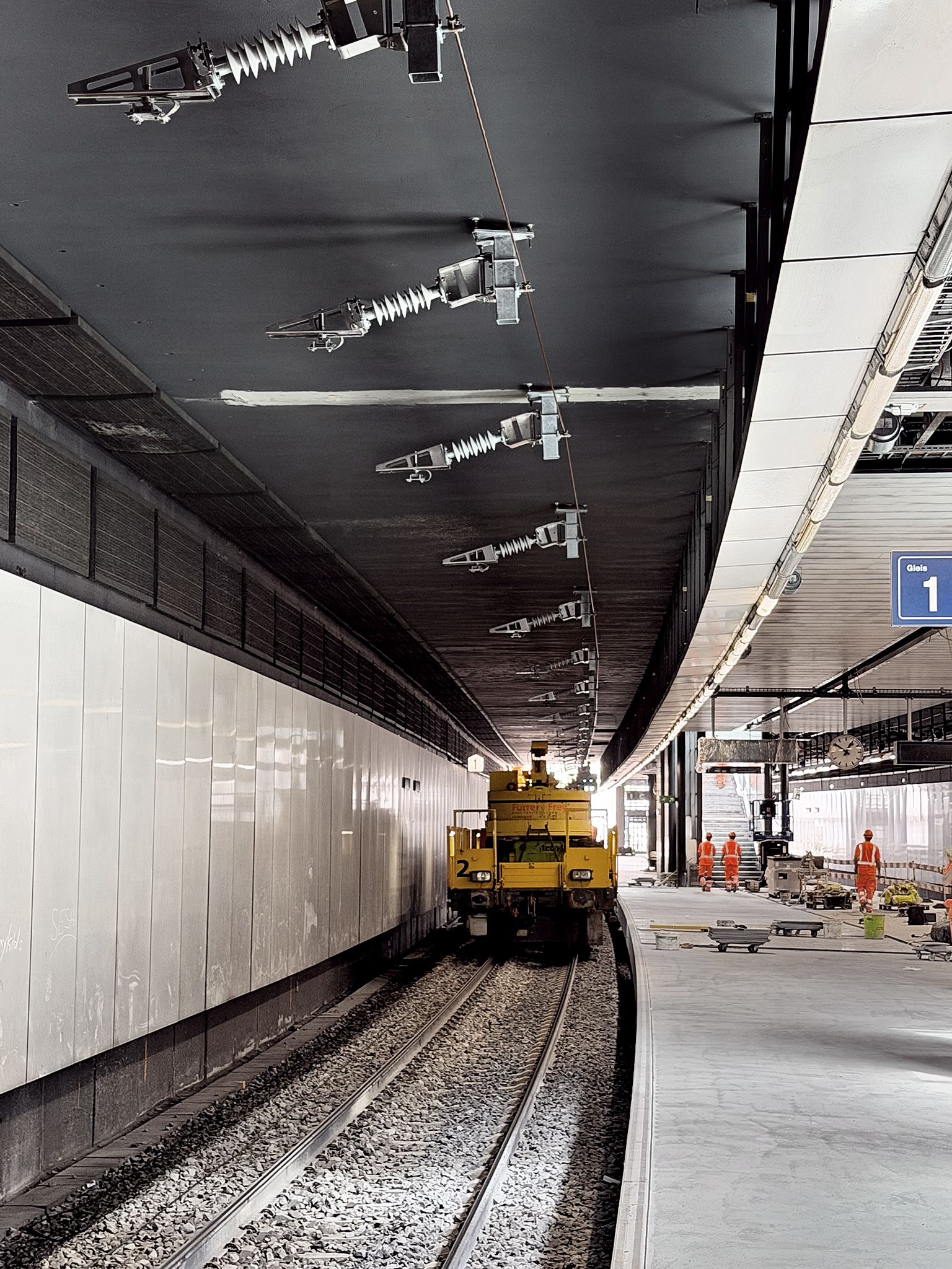 Ein gelbes Grossfahrzeug steht auf einem Gleis im Bahnhof Opfikon, und rechts davon stehen Bauarbeiter in orangefarbenen Westen auf dem Bahnsteig. Über dem Gleis sind Fahrleitungen zu sehen.