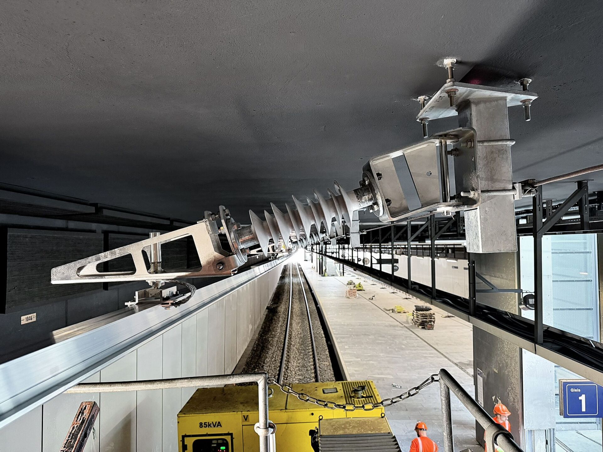 Im modernen Bahnhof Opfikon ist ein grossen DSS-Schwenktragwerk an einer Metallhalterung über den Gleisen angebracht; darunter sind auf dem Bahnsteig Arbeiter und Arbeitsgeräte zu sehen.