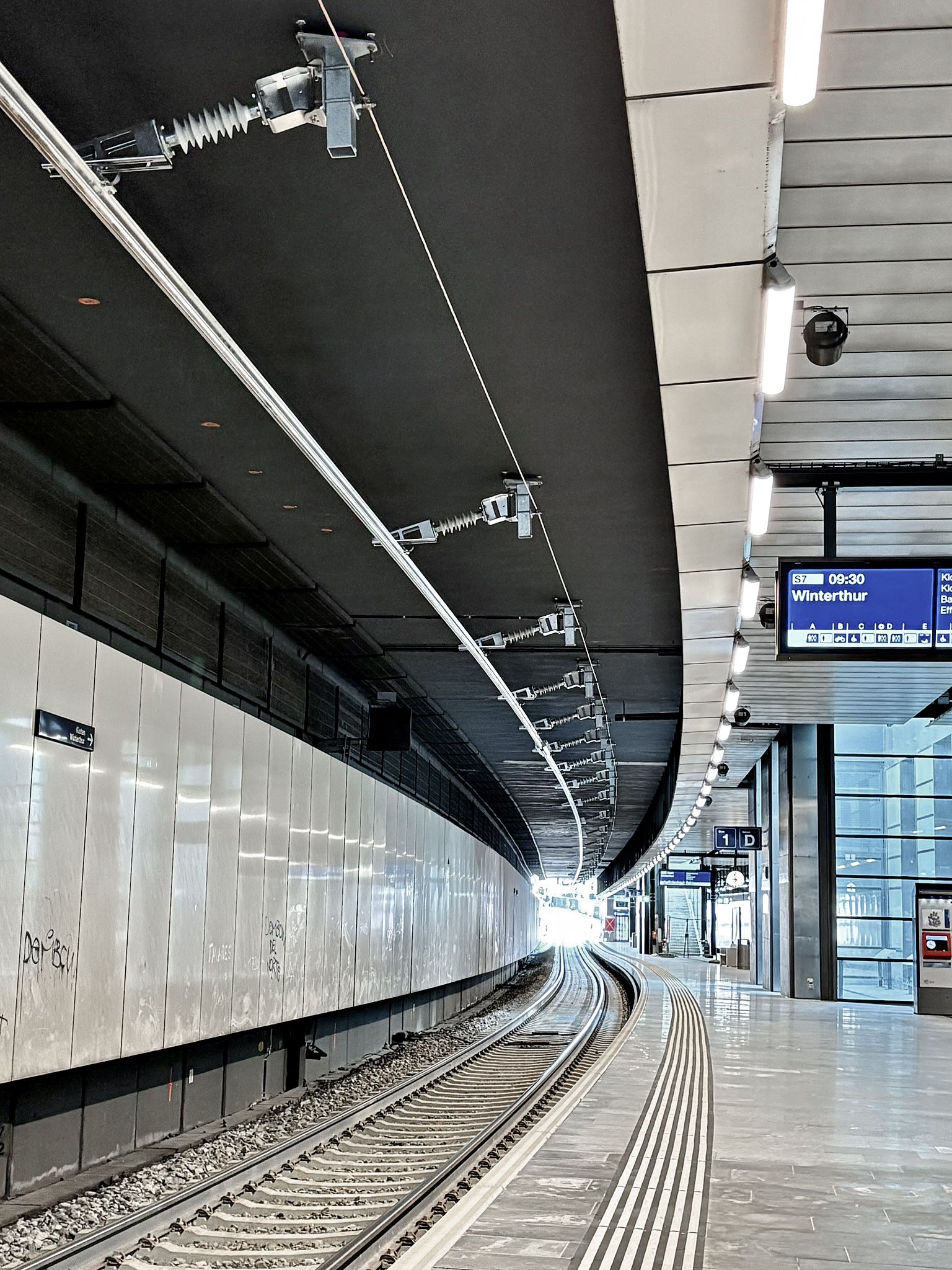 Ein moderner Bahnsteig am Bahnhof Opfikon mit geschwungenen Gleisen, Deckenbeleuchtung, Deckenstromschiene und einer digitalen Anzeige mit der Aufschrift „Winterthur“. Der Bereich ist sauber, mit glatten Wänden und einem weitgehend leeren Bahnsteig.