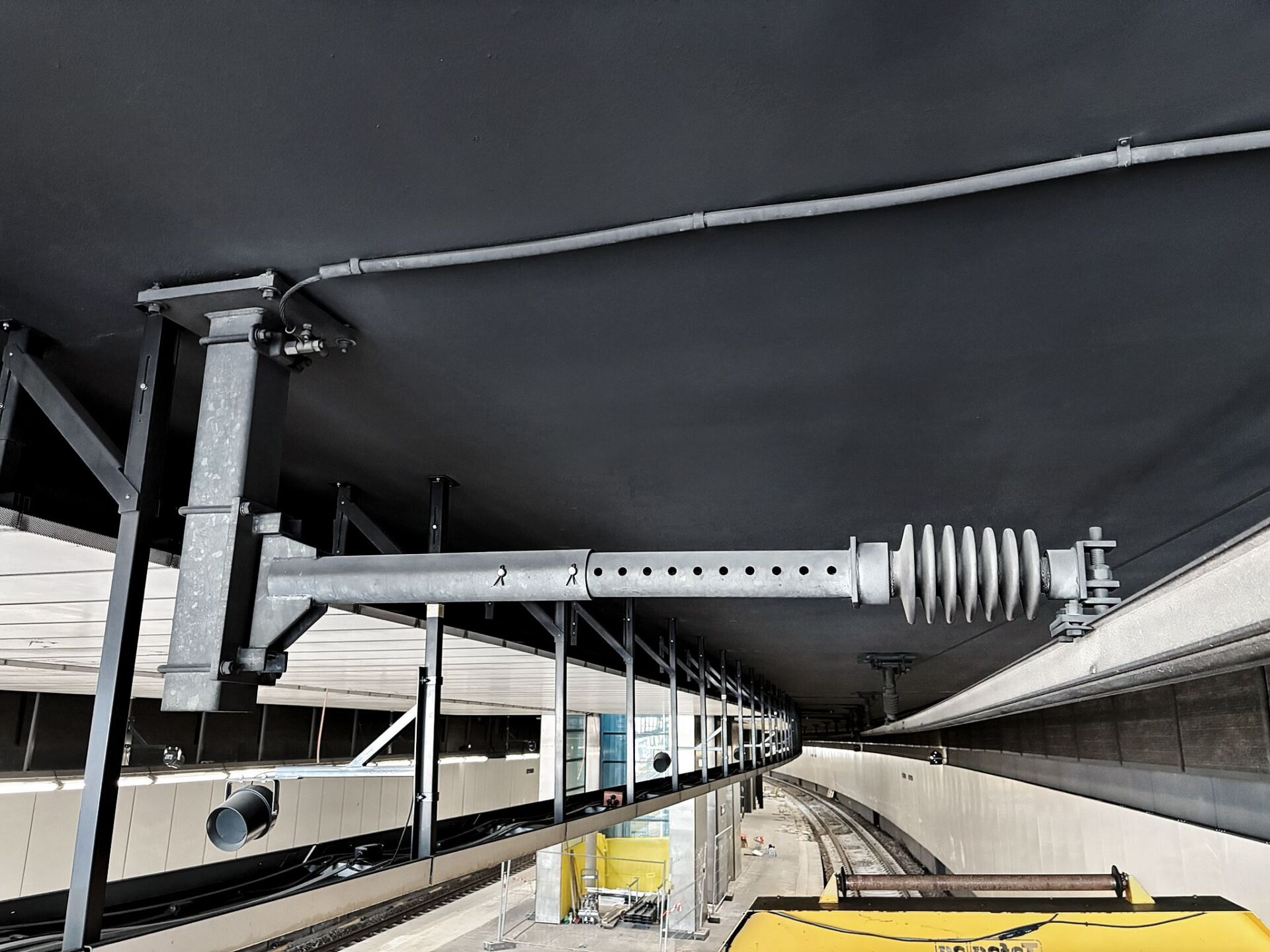 An einer schwarzen Decke über einem modernen Bahngleis ist ein DSS-Schwenktragwerk aus Metall angebracht; darunter sind Bauzäune und Kabel zu sehen.