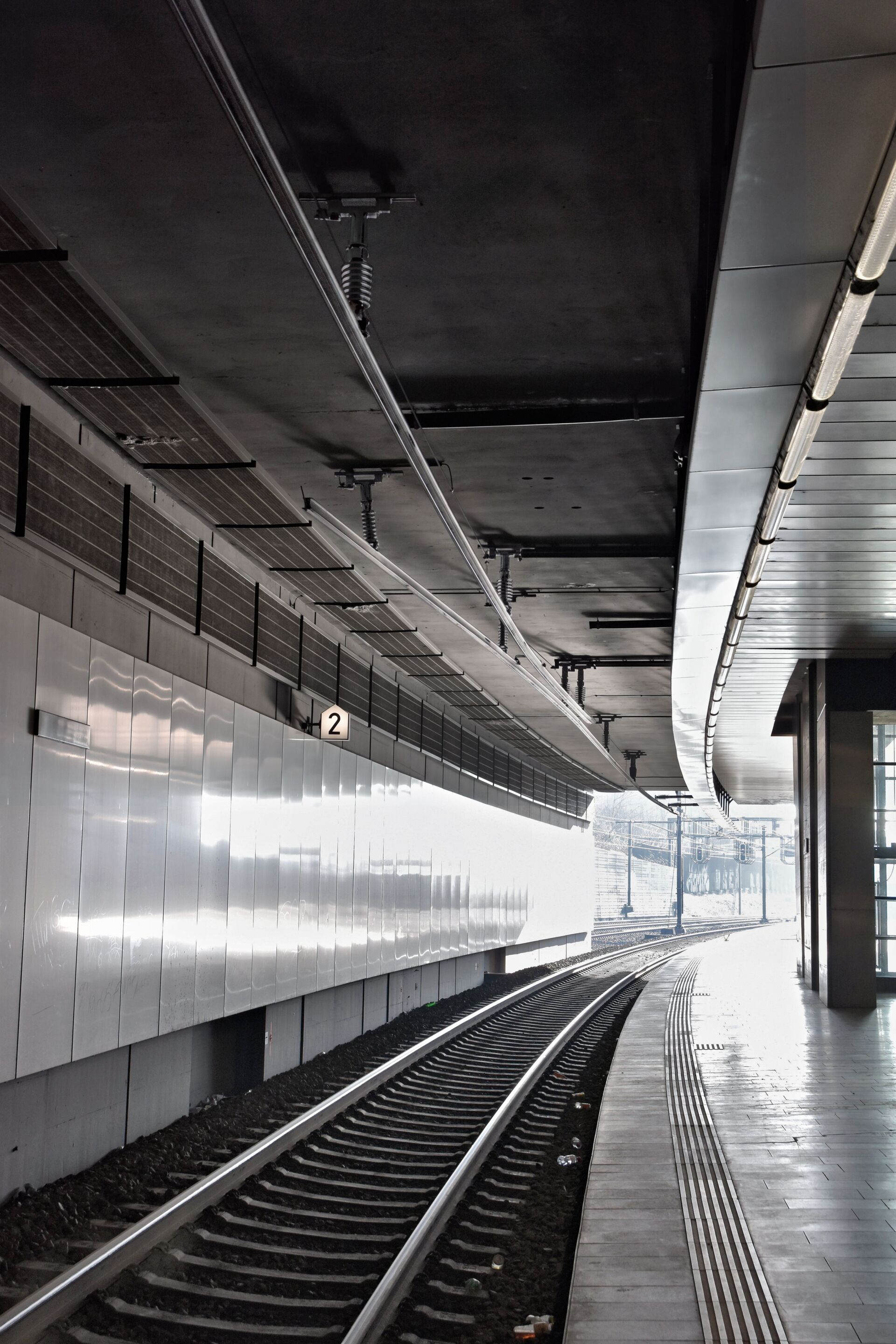 Leerer, moderner Bahnsteig mit geschwungenen Gleisen, Fliesenboden und einer weißen Wand. Deckenleuchten beleuchten den Bereich. Über dem Bahnsteig hängt ein Schild mit der Nummer 2.