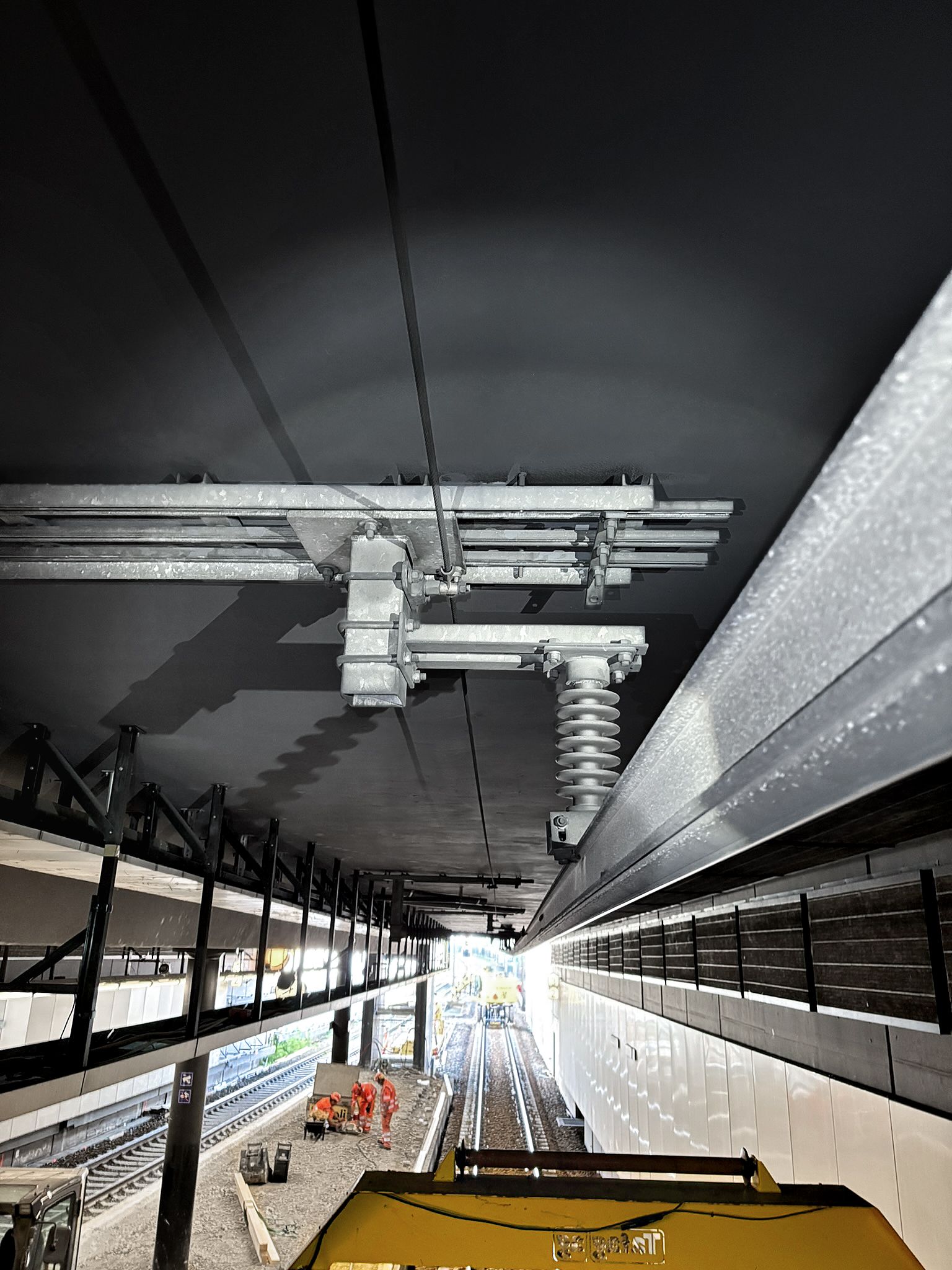 Blick auf einen im Bau befindlichen Bahnhof mit sichtbaren Fahrleitungen, Gleisen, Bauarbeitern und teilweise fertiggestellten Wänden. Die Szene ist hell ausgeleuchtet, wodurch bauliche Details hervorgehoben werden.